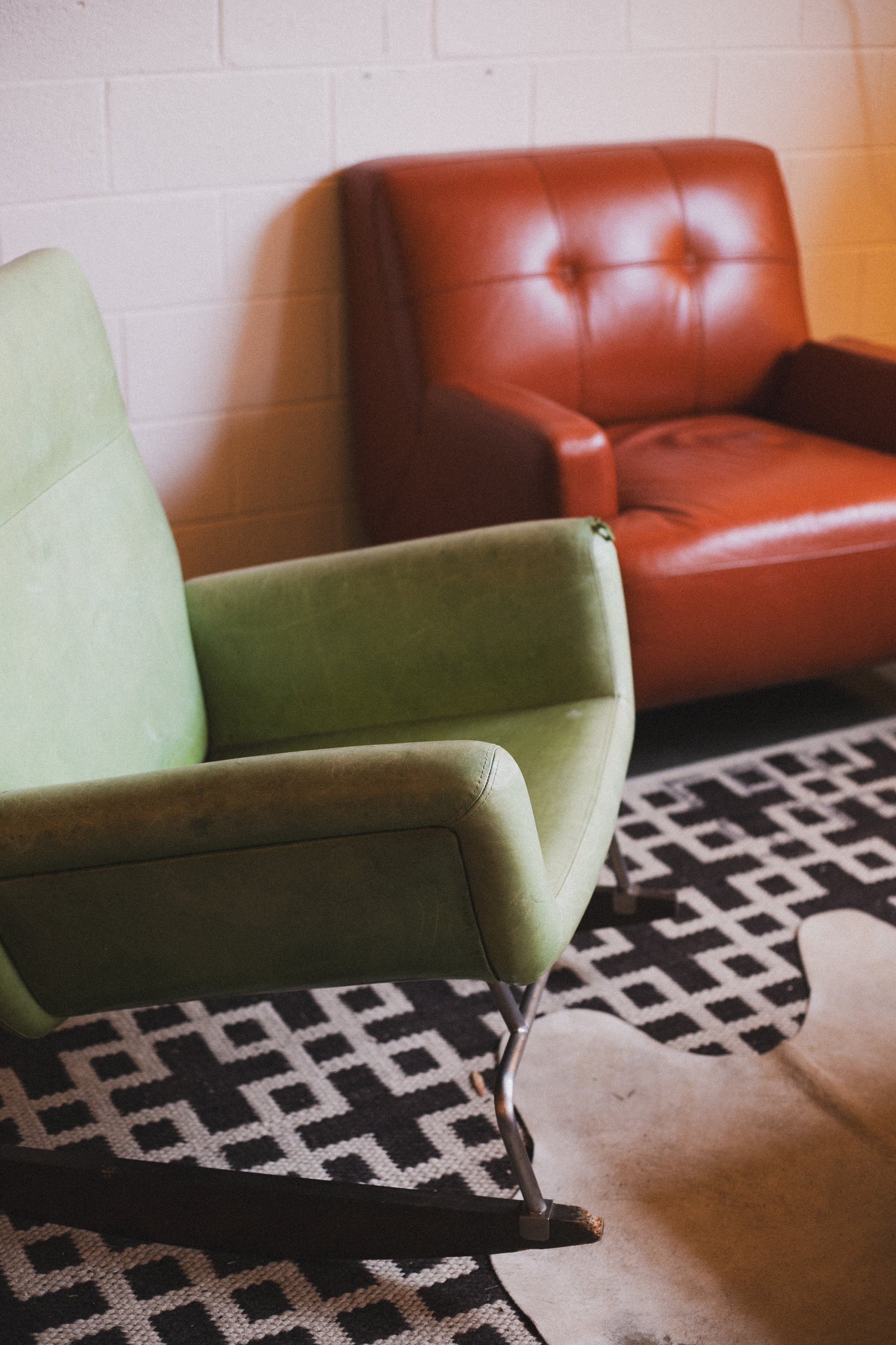 A green chair and a red leather armchair are positioned on a black and white patterned rug in front of a white brick wall.