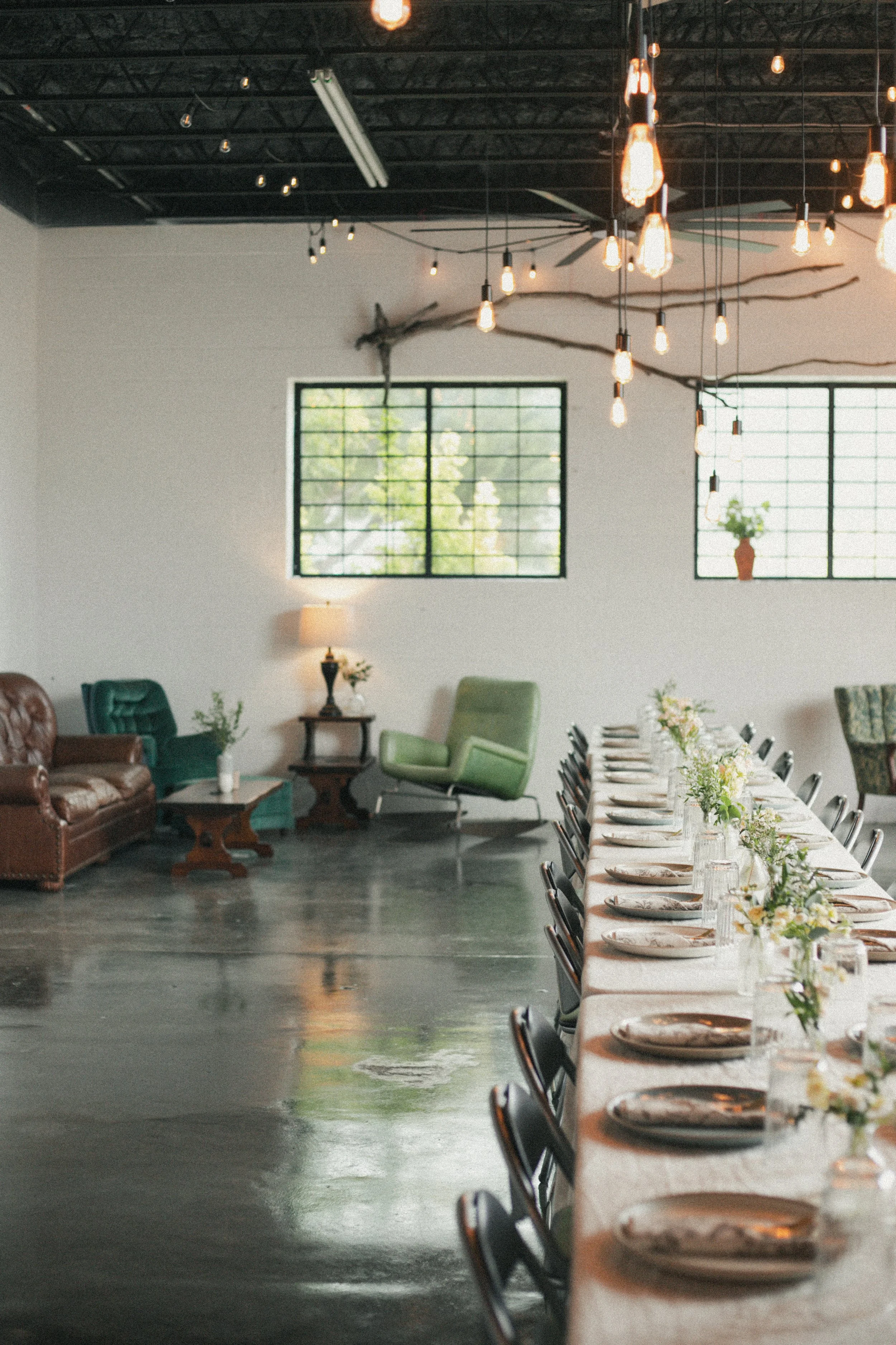 A long dining table set for a formal event with plates, glasses, and floral centerpieces, with black chairs arranged along both sides of the table. In the background, there are vintage lounge chairs, a side table with a lamp, and large windows letting in natural light. The room has a modern industrial style with a black ceiling, hanging Edison bulbs, and exposed beams.
