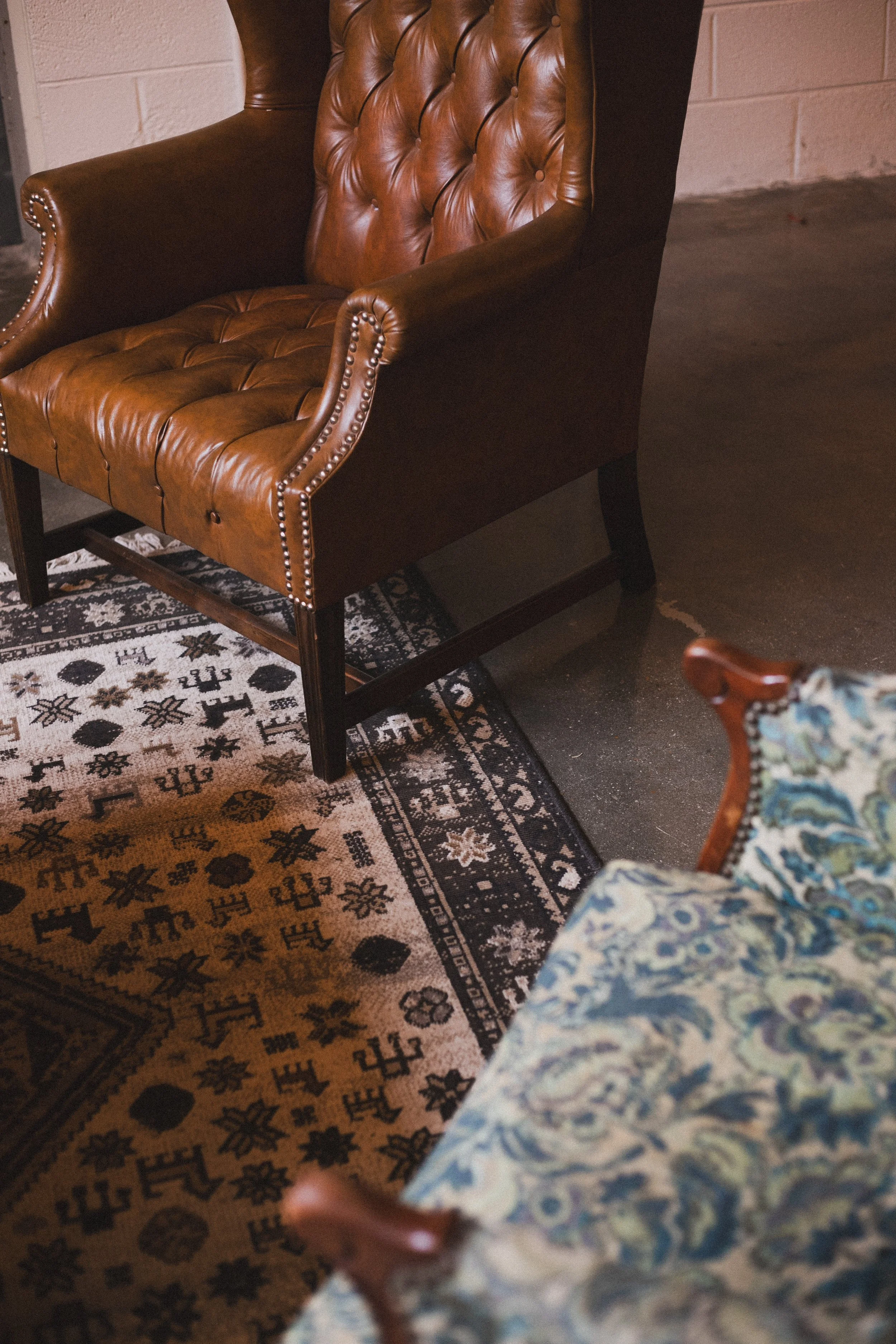 Close-up of a vintage brown leather armchair with tufted backrest and brass nailhead trim, placed on a patterned rug next to a fragment of a floral upholstered chair.