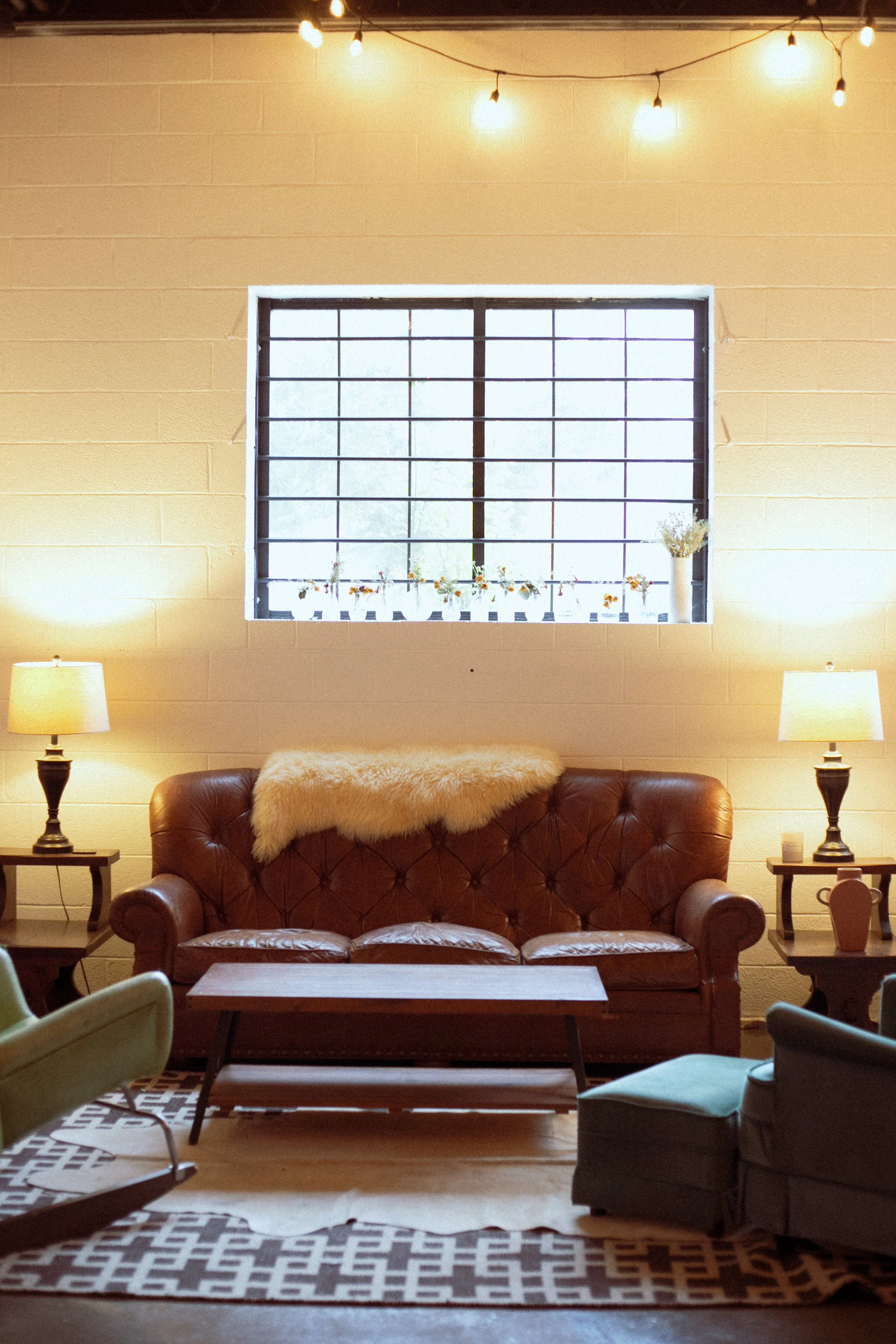 Living room with a brown leather tufted sofa, a light-colored fuzzy throw blanket, a wooden coffee table, two side tables with lamps, patterned rugs, a large window with metal bars, and string lights hanging from the ceiling.