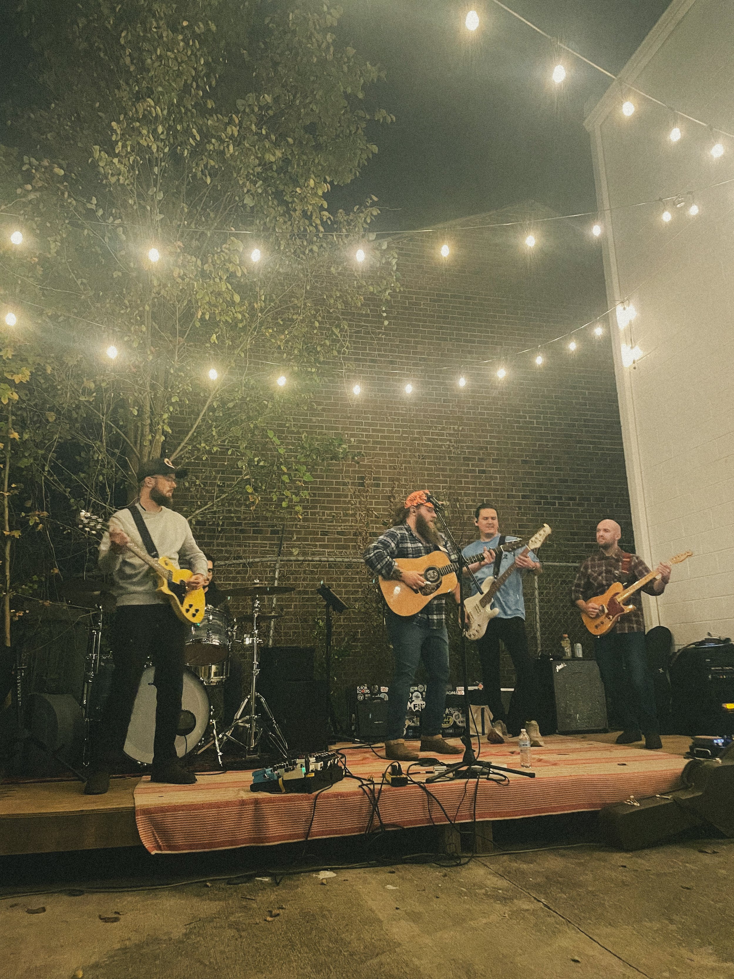 A band of five musicians performing on a stage outdoors at night, illuminated by string lights, with a brick wall and greenery in the background.