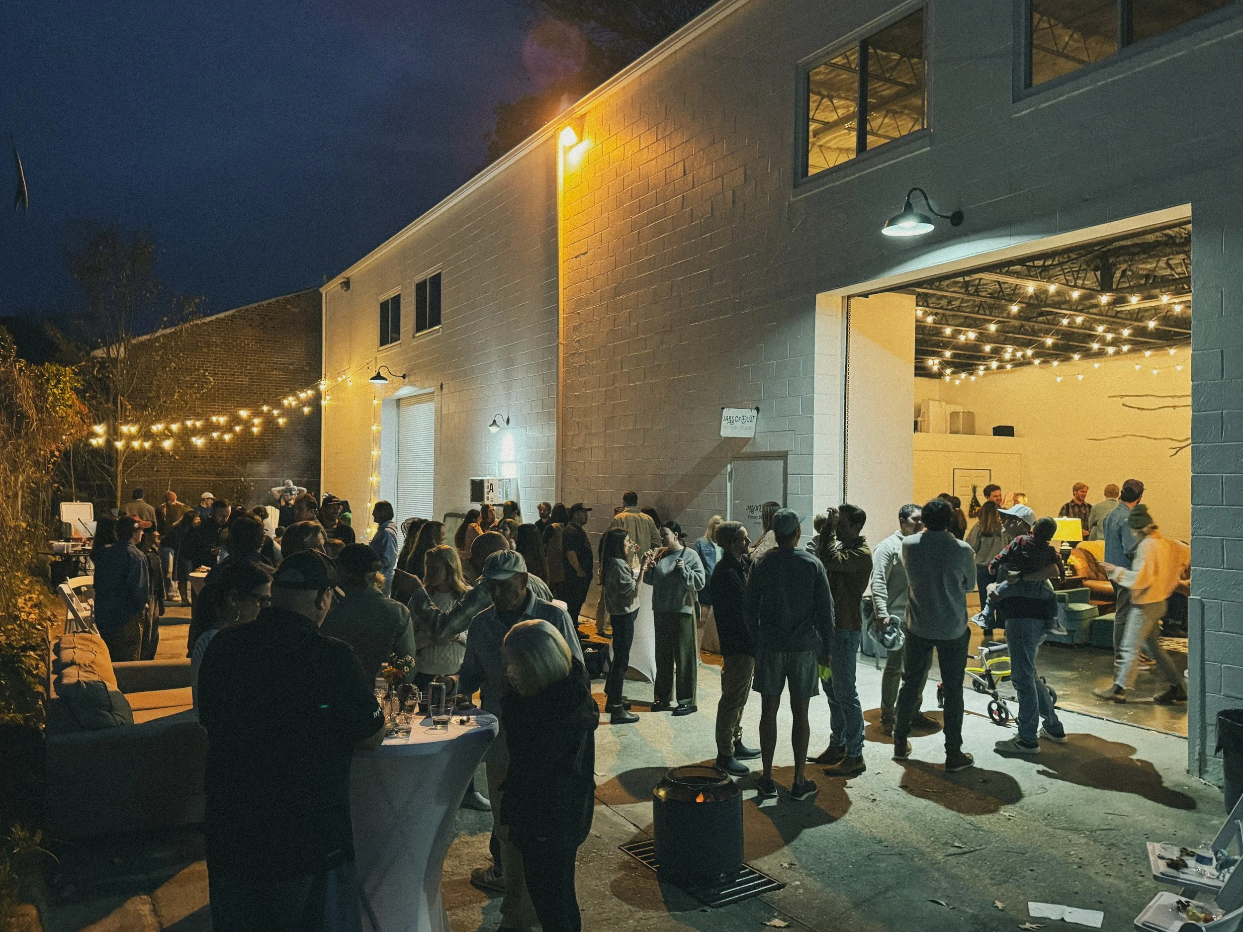 People gathered in an outdoor area at night, socializing and enjoying. The scene includes string lights, a building with open garage-style doors, and some tables with chairs, creating a festive atmosphere.