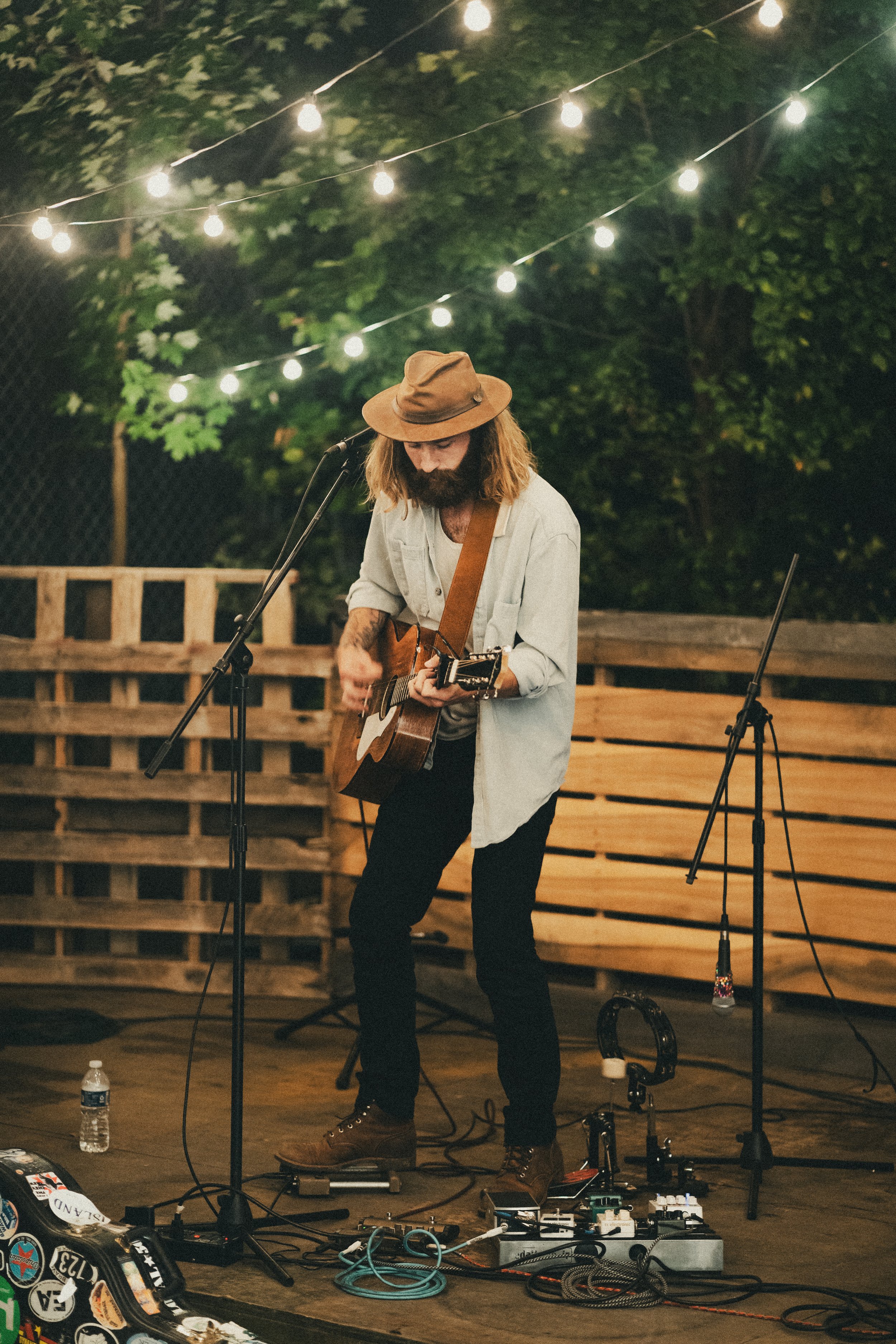 A man playing an acoustic guitar and singing into a microphone at an outdoor night event. He is wearing a tan hat, a light-colored shirt, and dark pants. The scene is decorated with string lights and has a backdrop of trees and a wooden pallet fence.