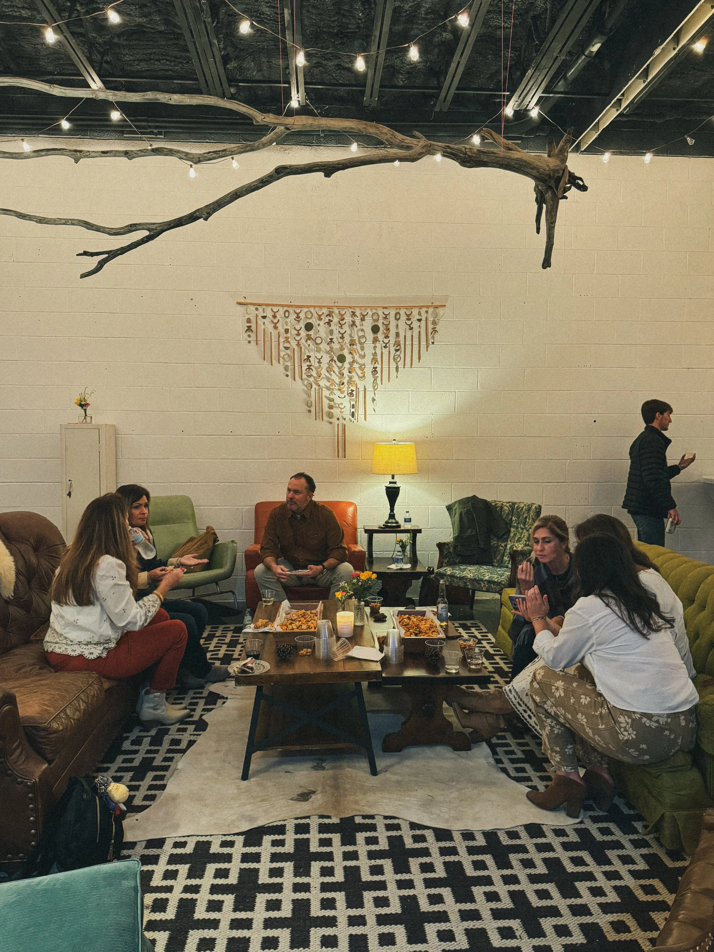 A cozy indoor gathering with people sitting on sofas and chairs around a low coffee table, enjoying food and drinks. The room has a beige brick wall decorated with a white and wooden hanging art piece, a small side table with a lamp, and string lights on the ceiling. The floor features a black and white geometric rug, and there is a large driftwood piece hanging from the ceiling.