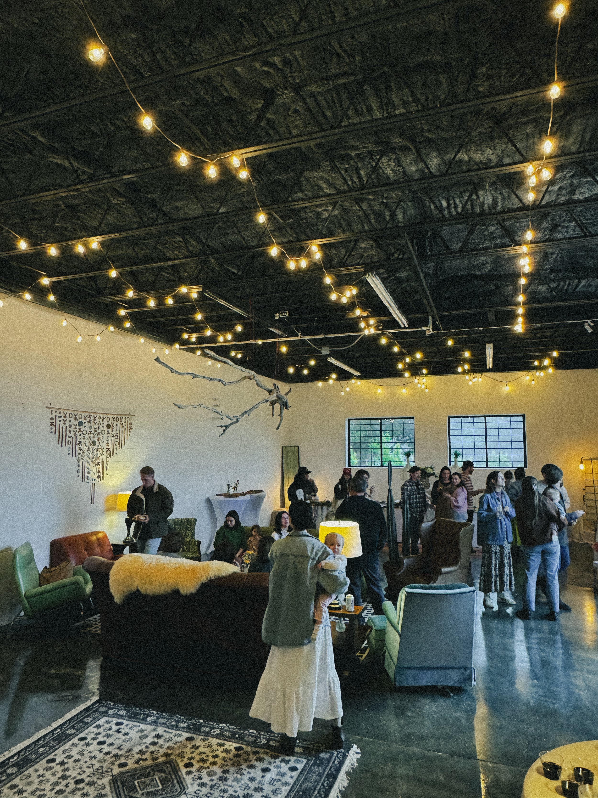 Indoor gathering at a modern, industrial-style space with string lights on the ceiling, people socializing and standing in groups, decorated with eclectic furniture including armchairs and a large patterned rug, a minimalist wall art piece, and a win