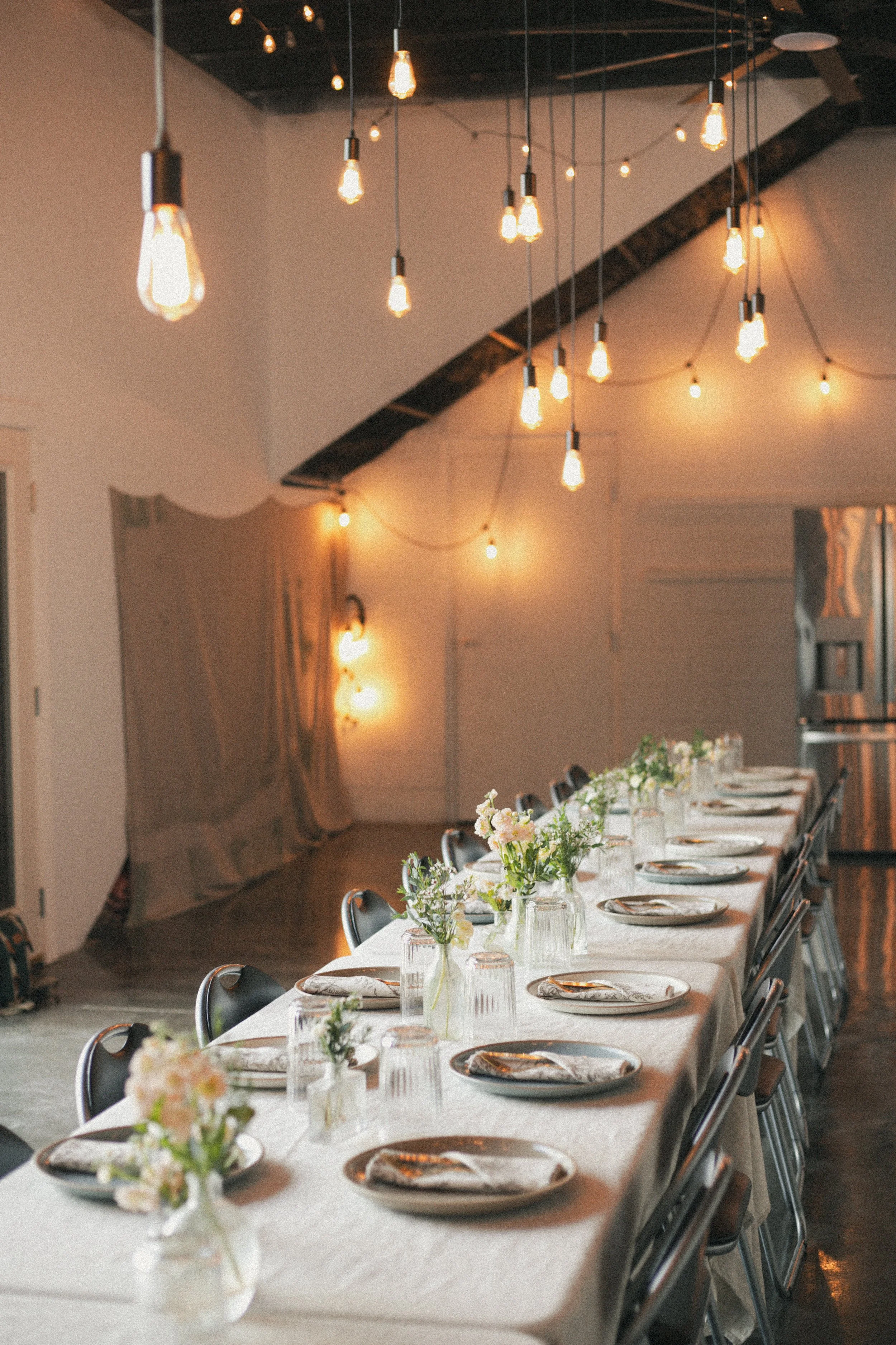 Interior of a dining area with a long table set with plates, napkins, and flower centerpieces. Hanging Edison light bulbs provide warm lighting. The room has a rustic style with wooden beams and curtains.