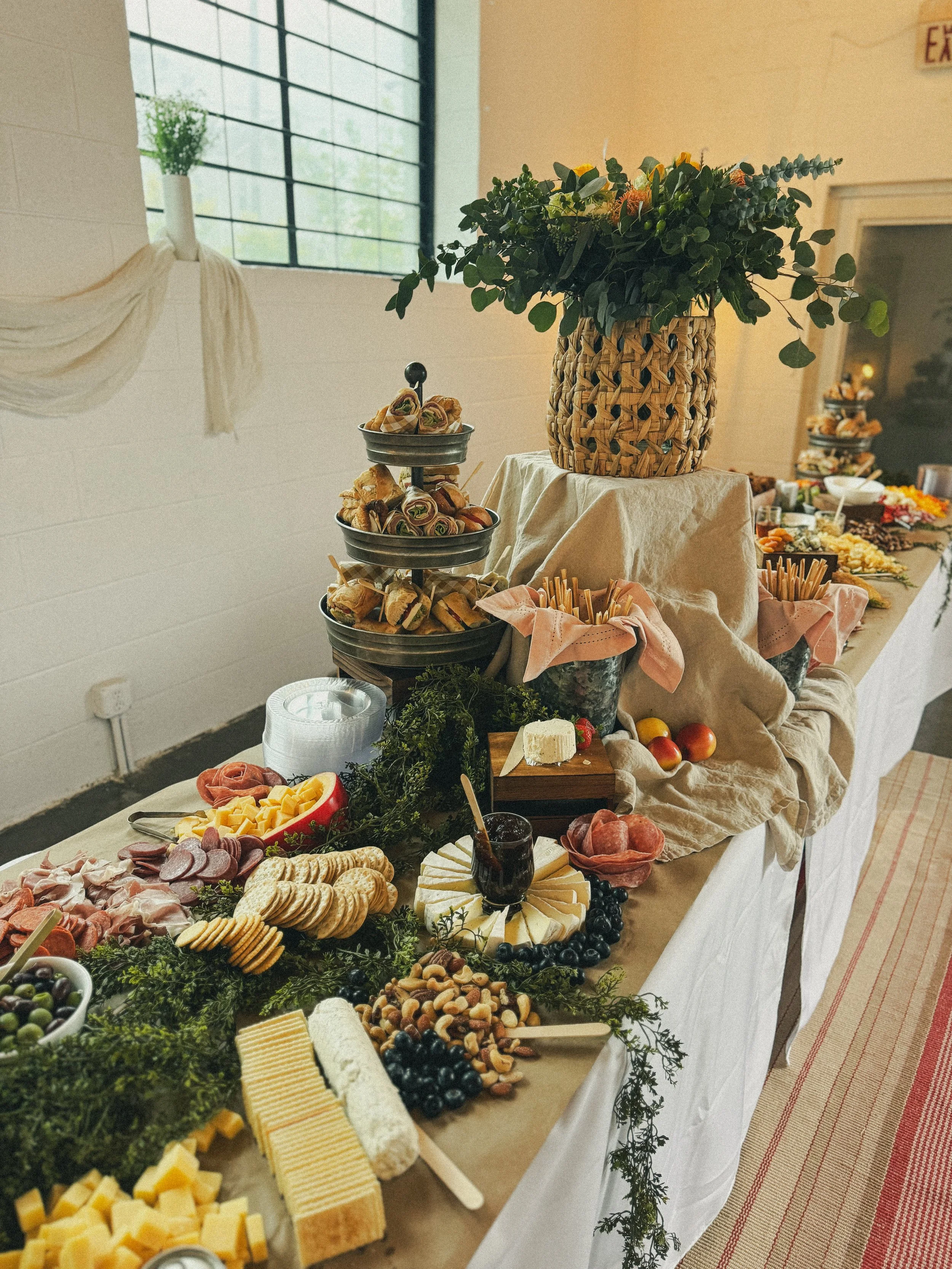 Table set for an event with various cheeses, meats, and snacks, decorated with greenery, a large woven basket with a floral arrangement, and serving trays stacked with deli items and fruits.
