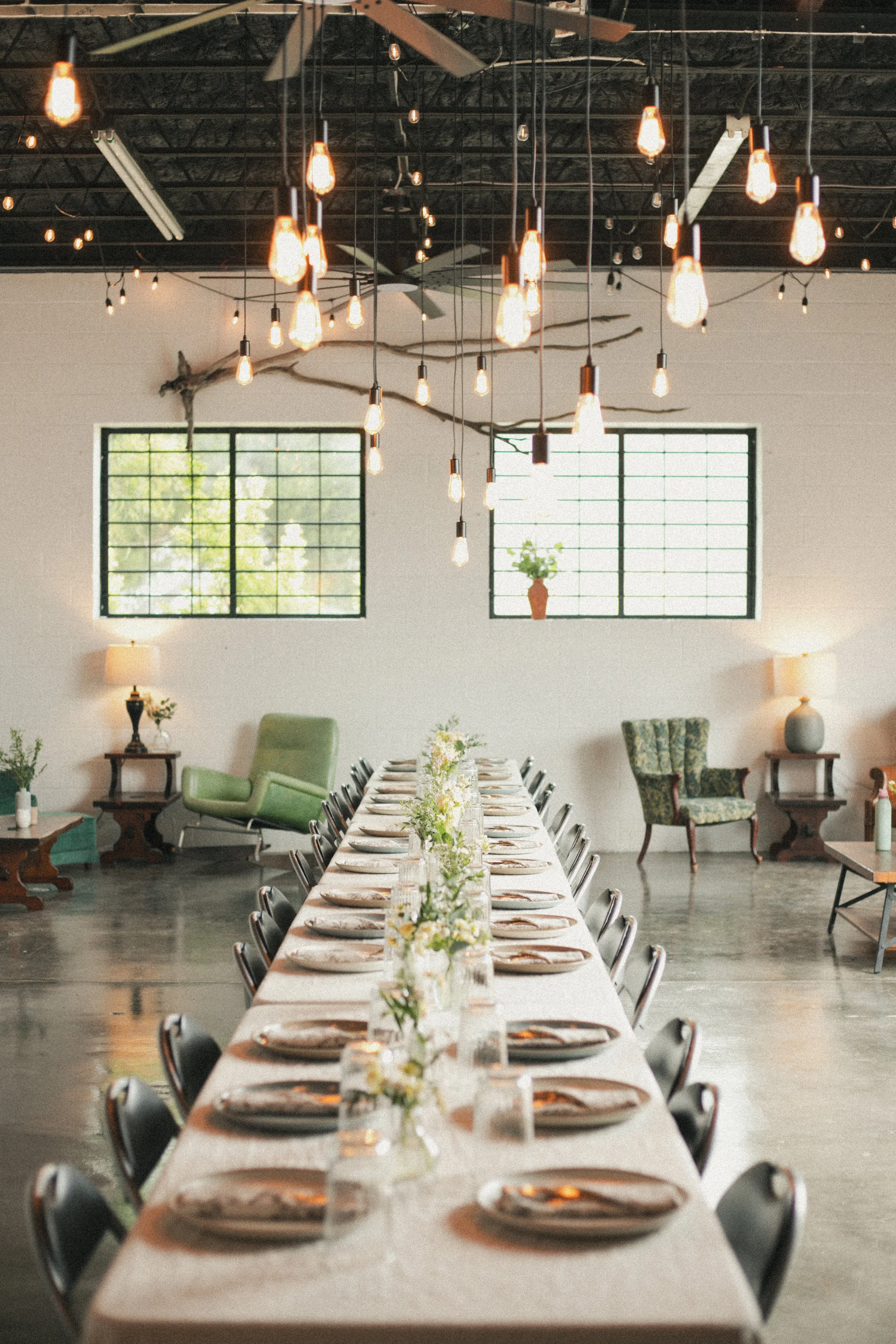 A long dining table set with plates, silverware, and small flower arrangements, in a modern, rustic-style room with large windows, green chairs, and exposed ceiling beams, illuminated by hanging Edison light bulbs.