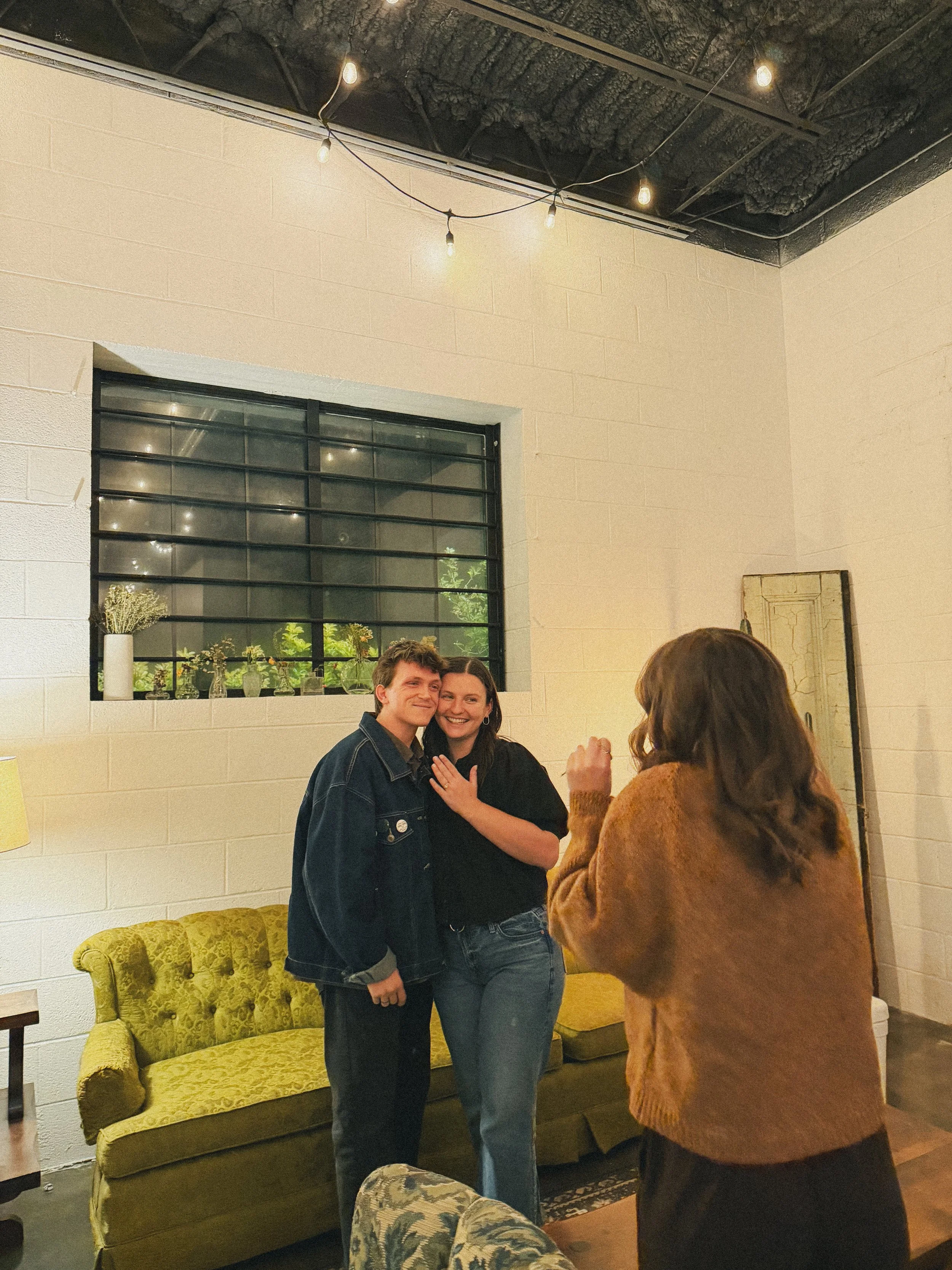 Two people are smiling and posing together for a photo, while a woman with brown hair in a brown sweater is taking a picture in a cozy indoor setting. The background features a white brick wall, a window with black metal bars, a yellow velvet sofa, and a mug sitting on a side table. There are string lights on the ceiling.