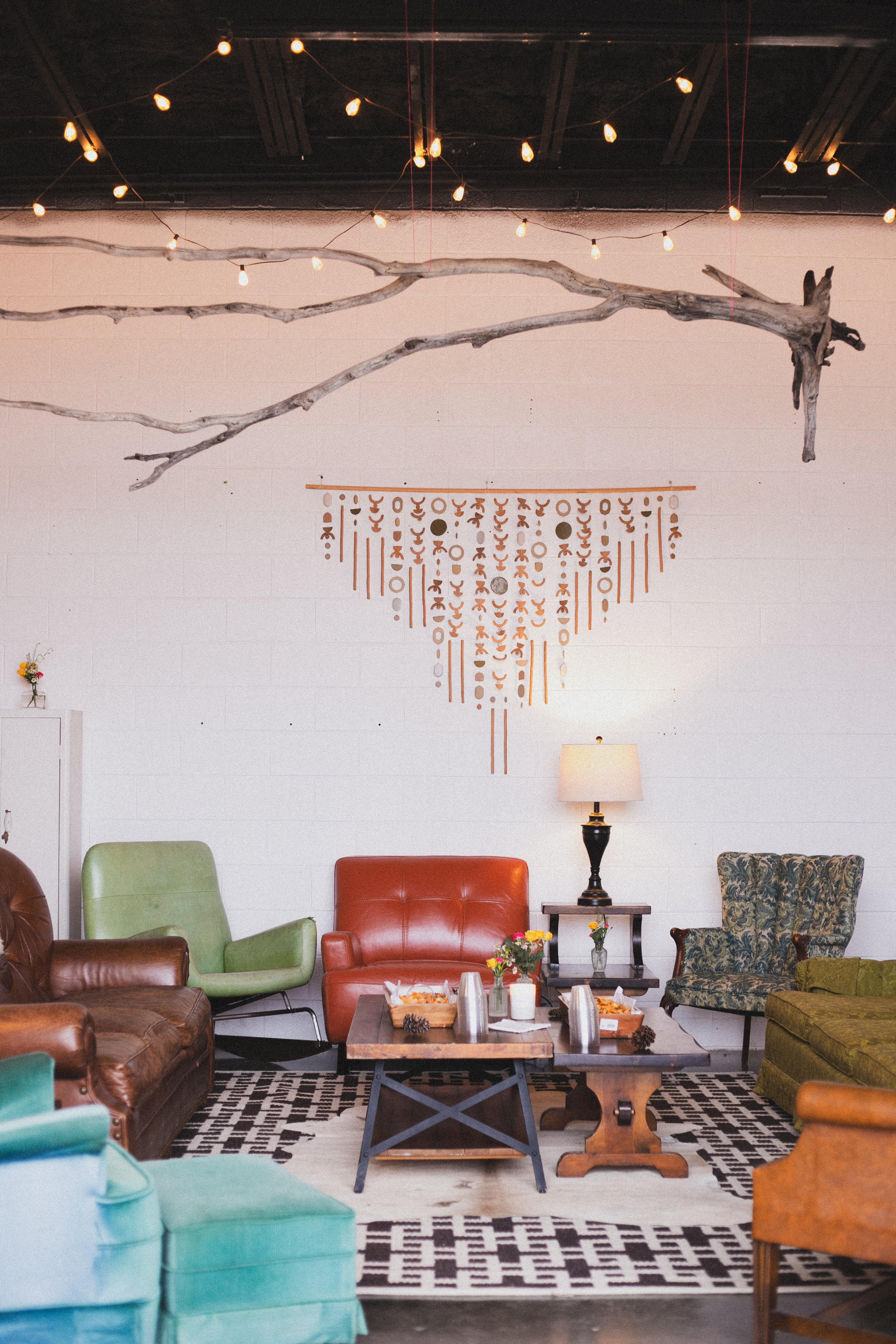 A cozy seating area with a variety of colorful chairs, a wooden coffee table with snacks and drinks, a patterned rug, a lamp, and decorative wall hanging.