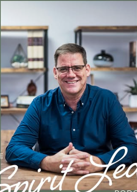 Smiling man wearing glasses and a blue shirt sitting at a table with hands folded, in a room with bookshelves and decor in the background.