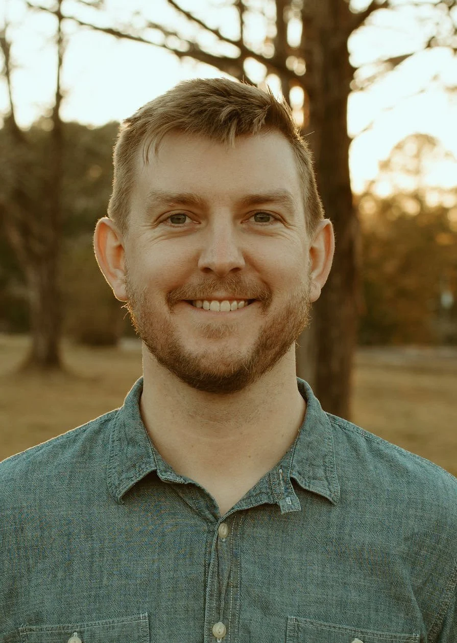 A smiling young man with short brown hair and a beard, wearing a denim shirt, standing outdoors during sunset with trees in the background.