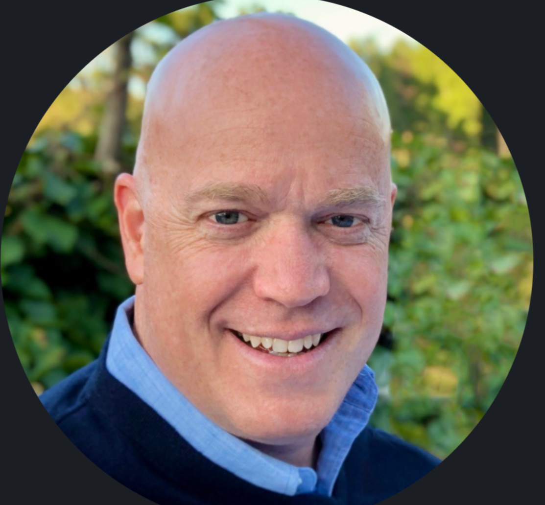 Headshot of a smiling, bald man with blue eyes, wearing a collared shirt and a dark sweater, outdoors with green foliage background.