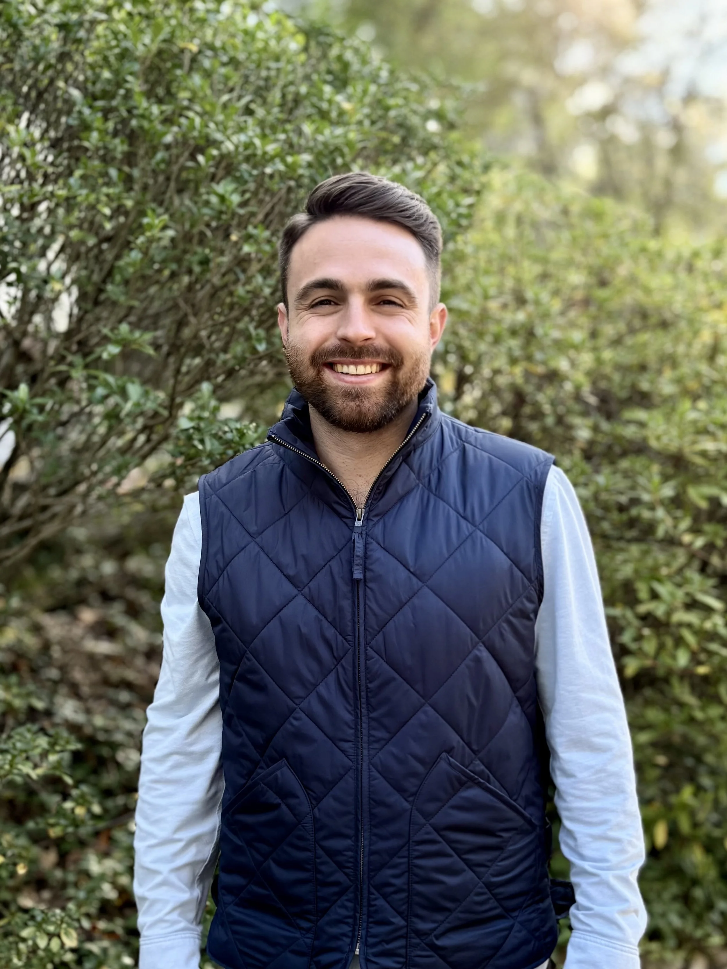 A smiling young man with a beard, wearing a navy blue quilted vest over a light gray long-sleeve shirt, standing outdoors with green shrubbery and trees in the background during daytime.