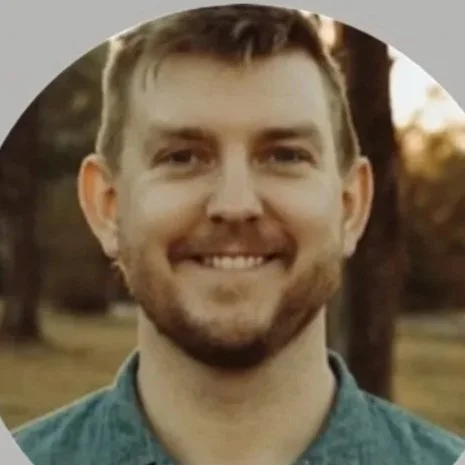 Young man with short brown hair and a beard smiling outdoors during sunset.