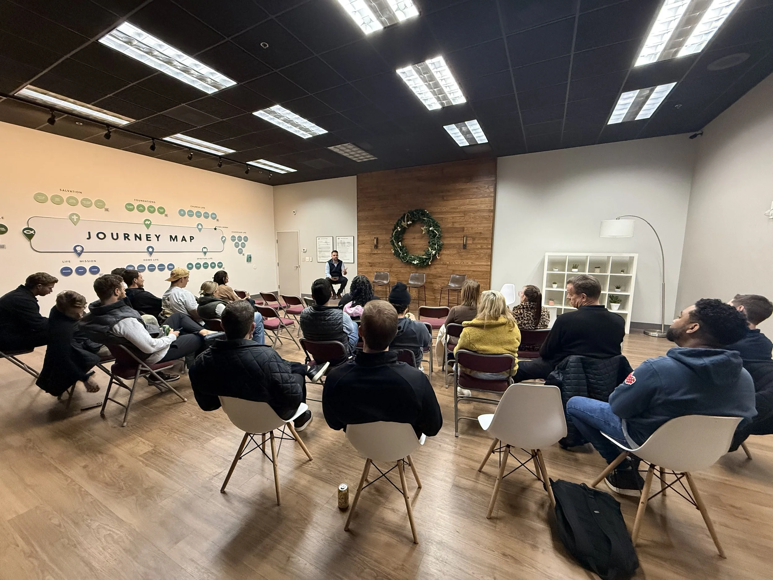 Group of people attending a presentation in a room with a wooden wall, wreath, and a journey map on the wall.