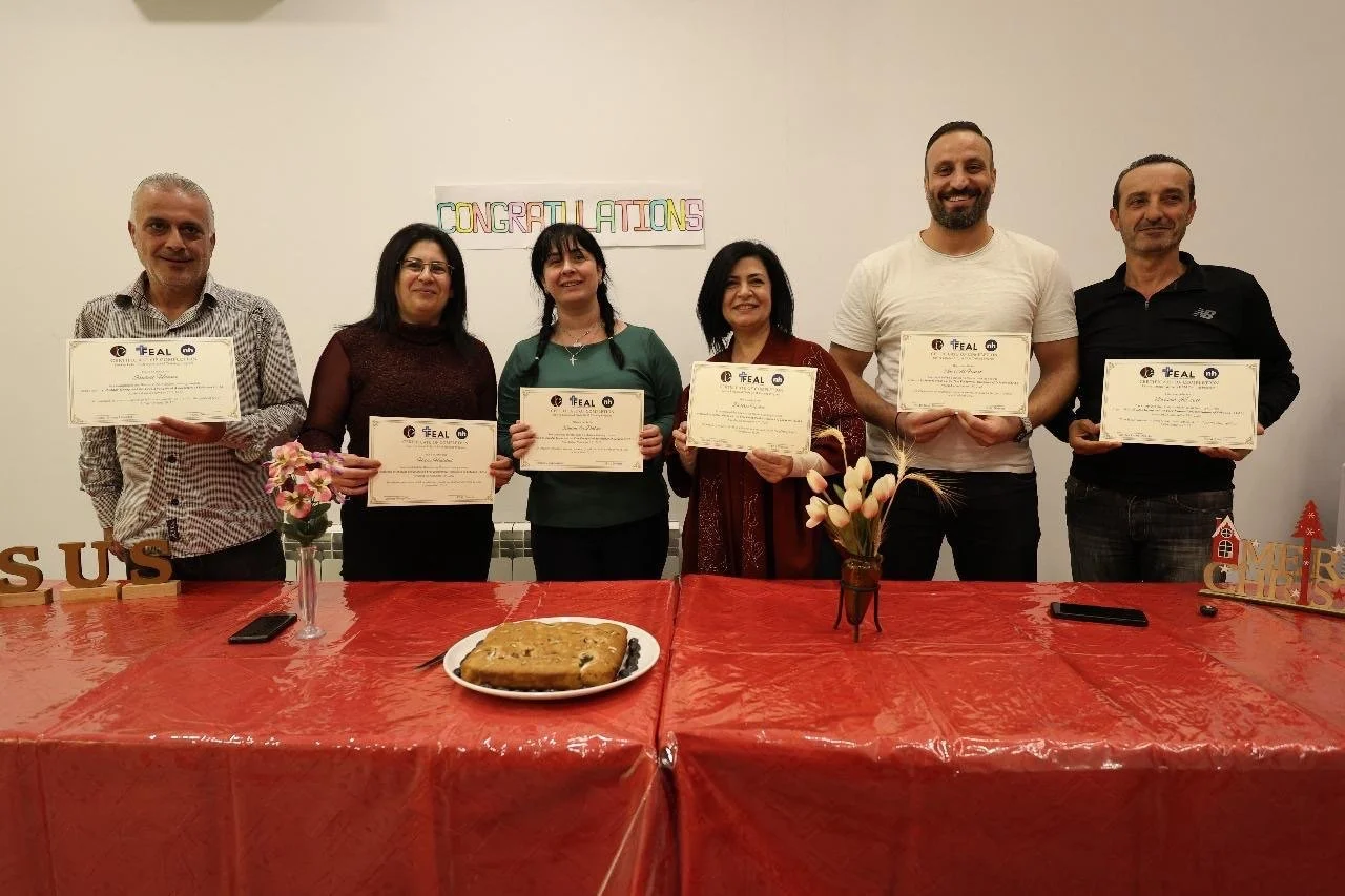 Six people standing behind a table holding certificates, with a sign reading 'CONGRATULATIONS' in the background, a cake, and decorations.