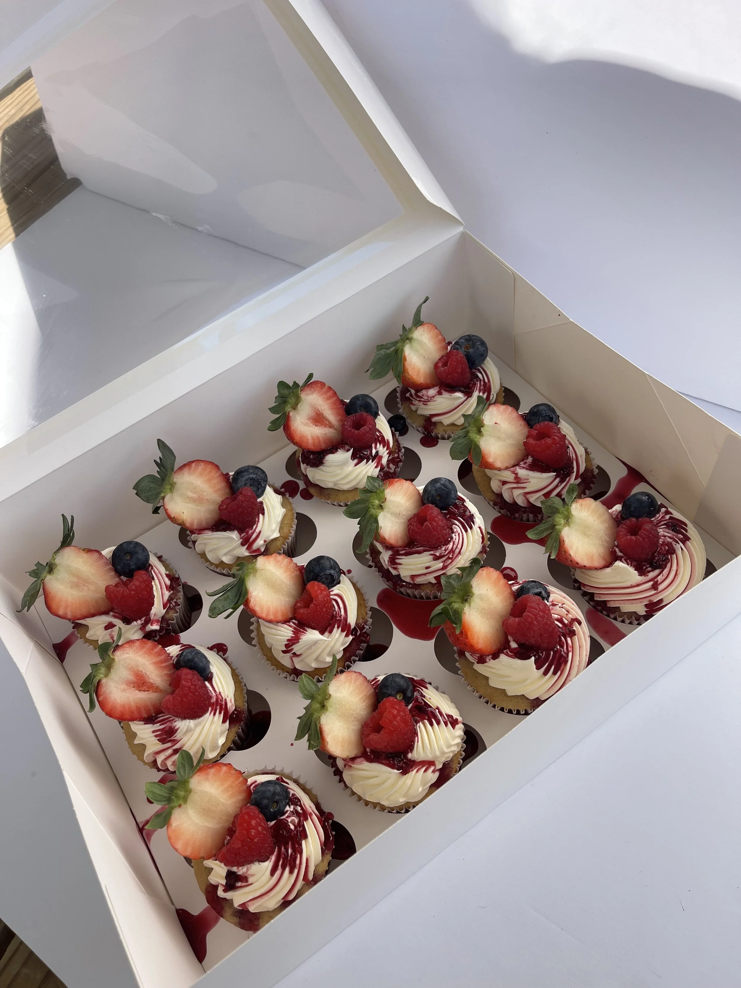 A white box filled with twelve mini fruit-topped cheesecakes, each decorated with strawberries, blueberries, and raspberries, with some fruit drizzled with red syrup.