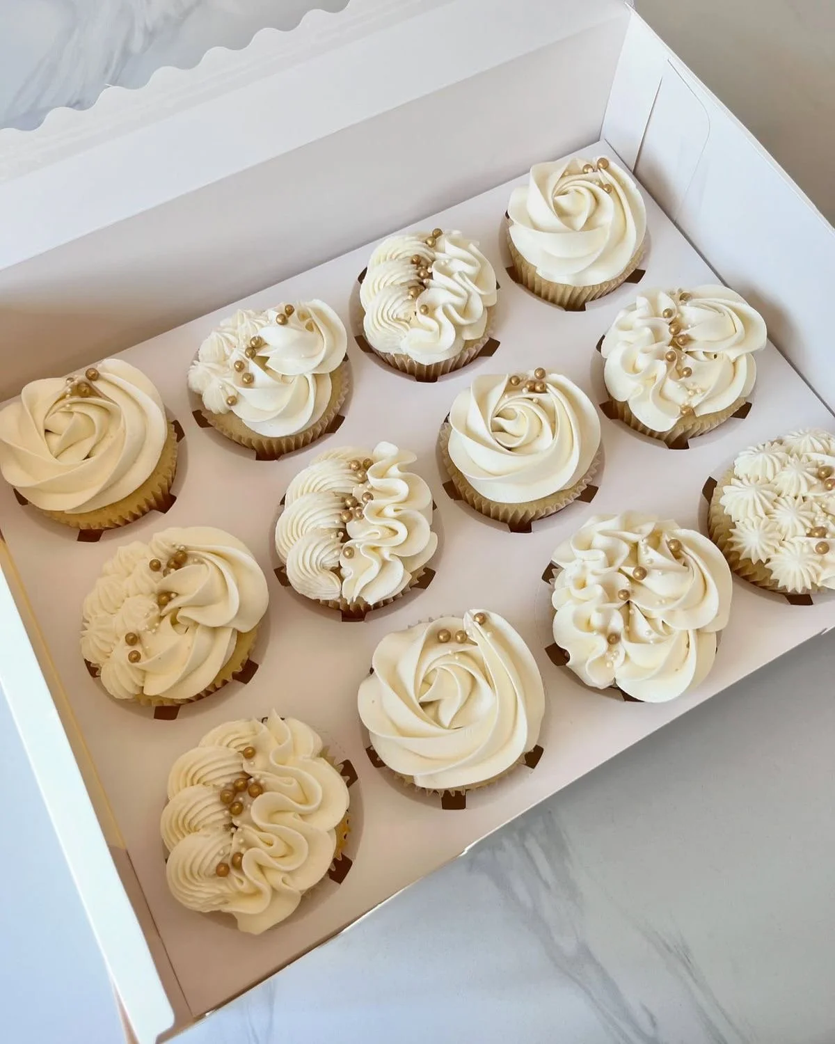 A white bakery box containing twelve decorated cupcakes with white frosting and gold decorative pearls.