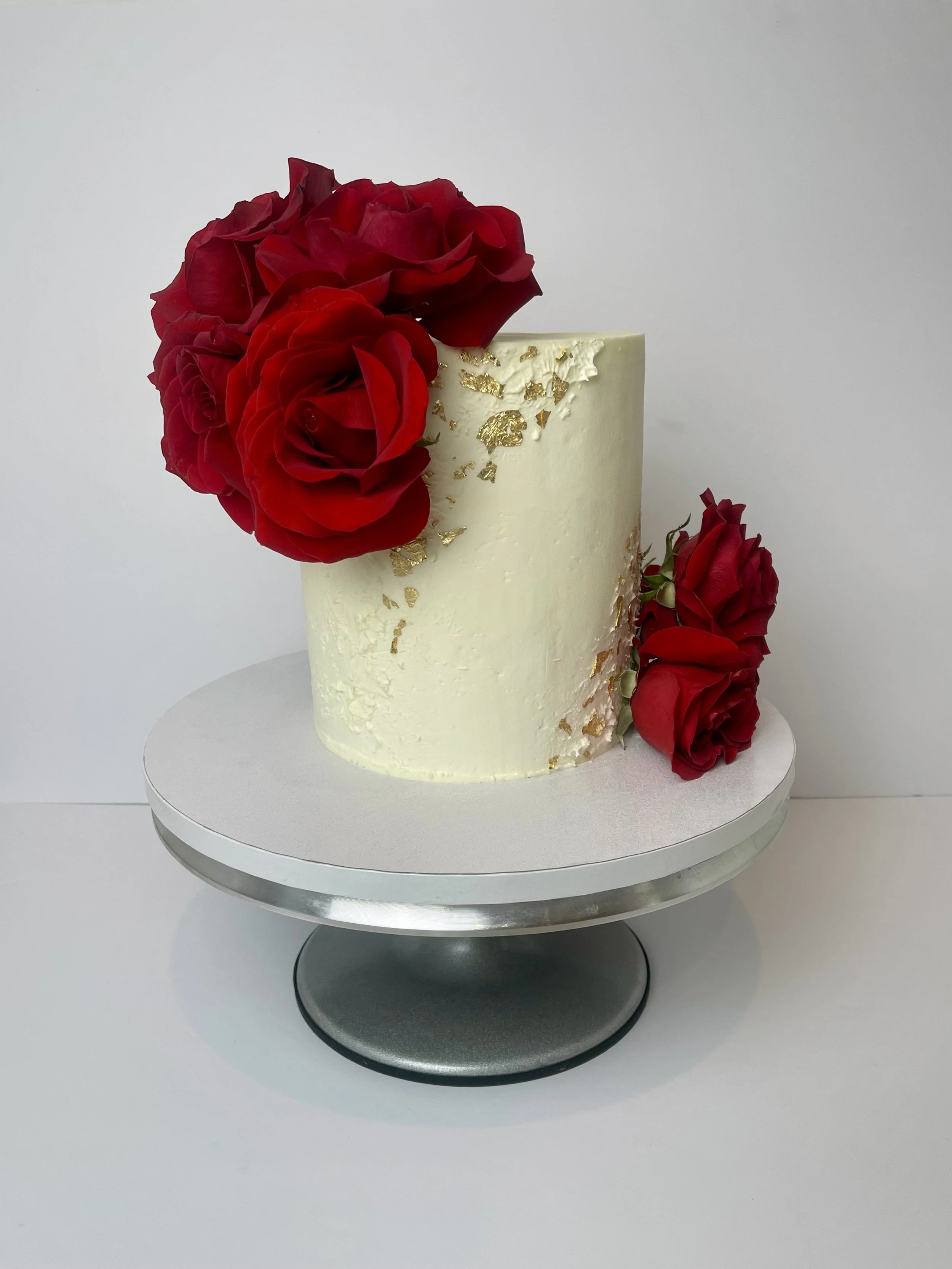 A white cylindrical cake decorated with red roses and gold flakes, placed on a silver cake stand.
