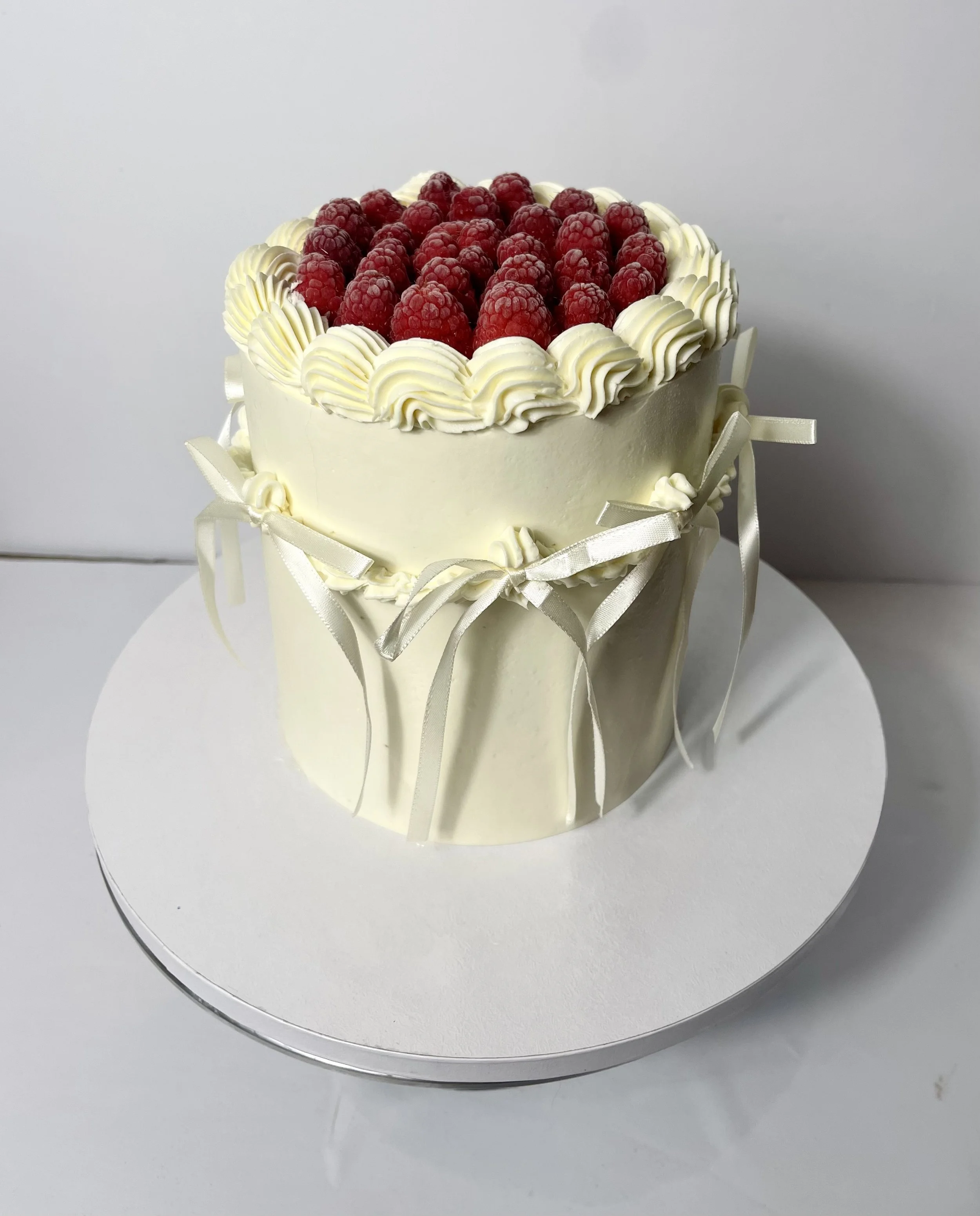 White anniversary cake decorated with piped cream and tied with satin ribbons, topped with fresh raspberries, on a white cake board.