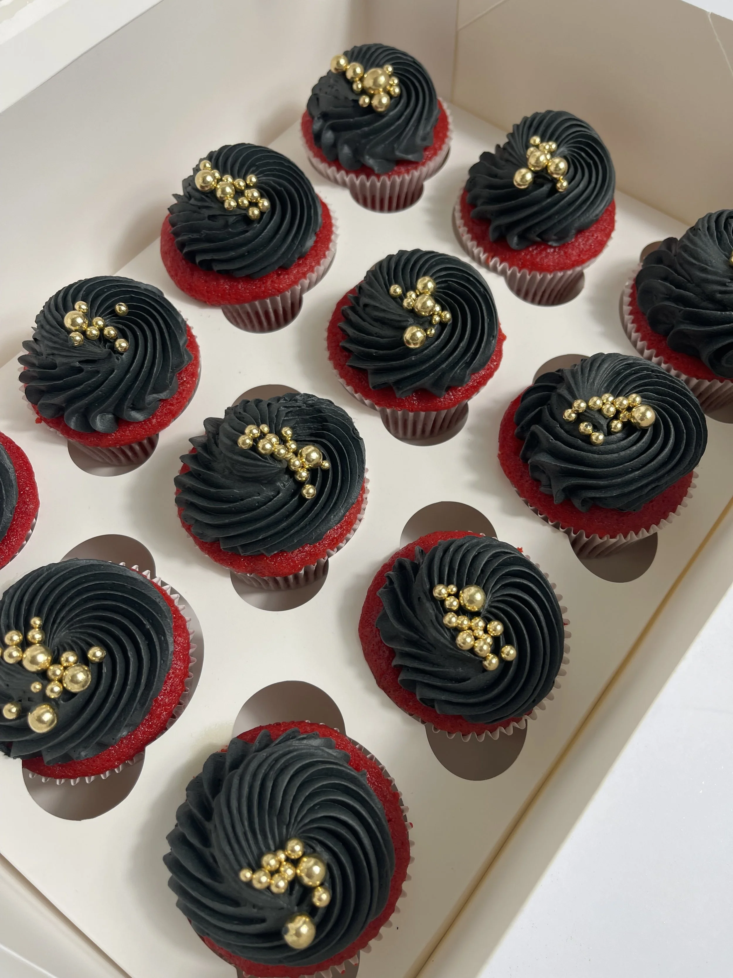 Red velvet cupcakes with black frosting and gold decorative pearls arranged in themed swirls on a white tray.