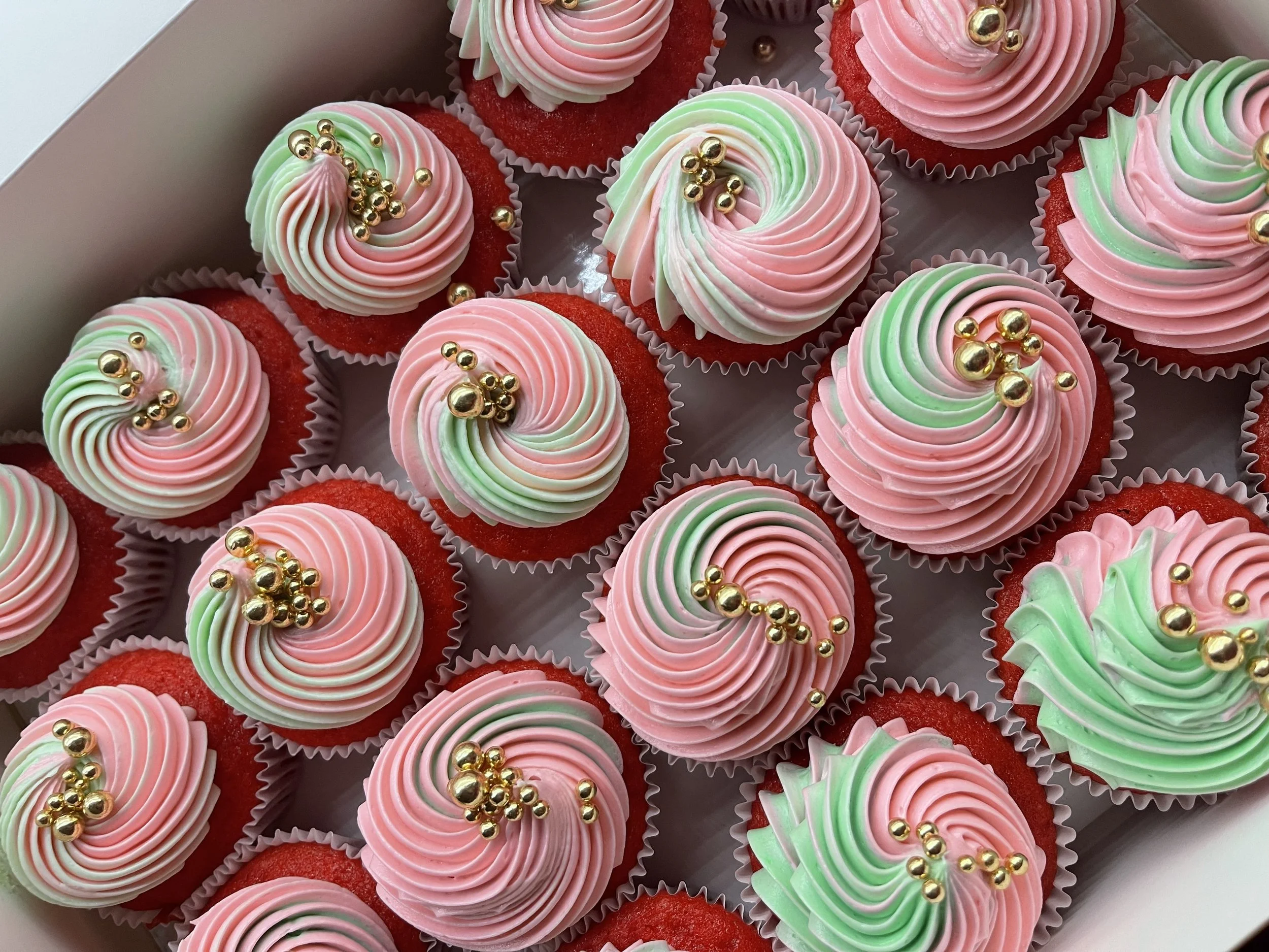 Decorated red velvet cupcakes with pink, white, and green swirl frosting topped with gold edible beads in a white box.