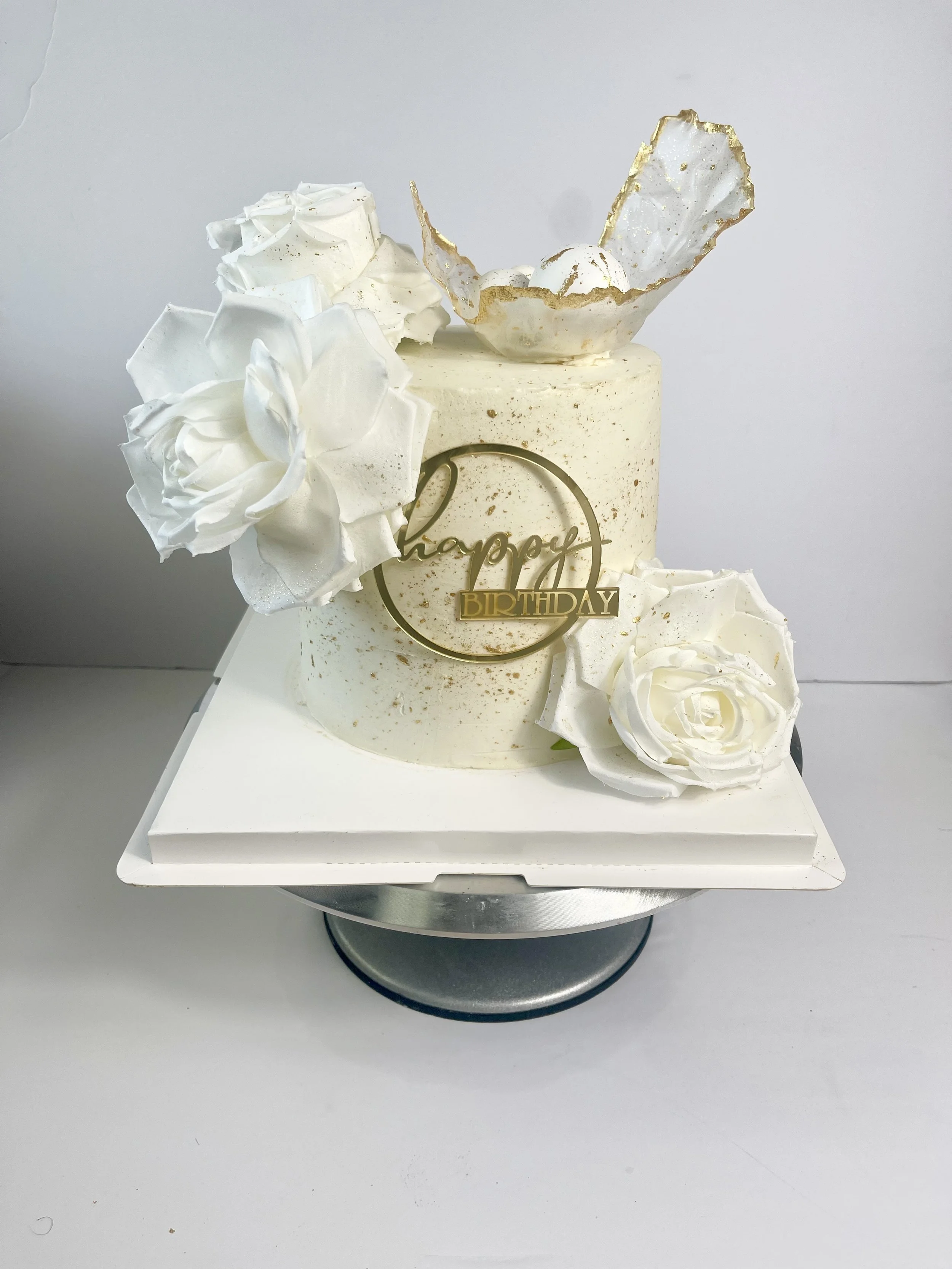 White birthday cake with white floral decorations and a gold 'happy birthday' topper, placed on a silver cake stand.