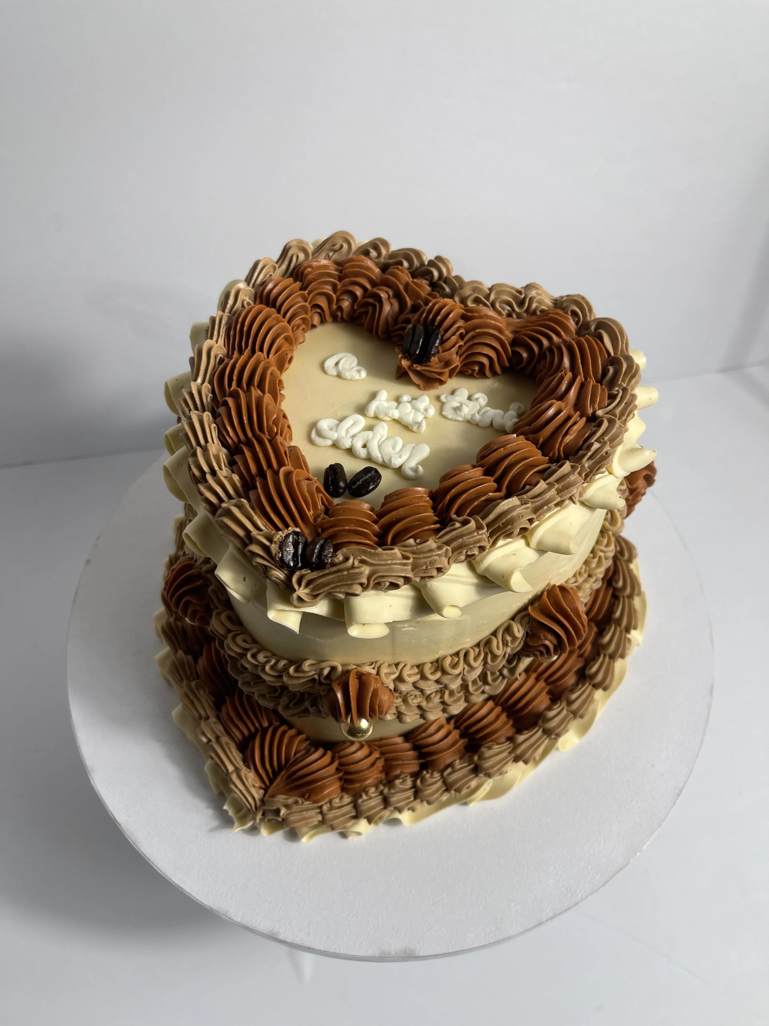 Heart-shaped, cream-colored cake decorated with various shades of chocolate frosting and coffee beans. The top has a message written in white frosting that reads "Happy Eddle."