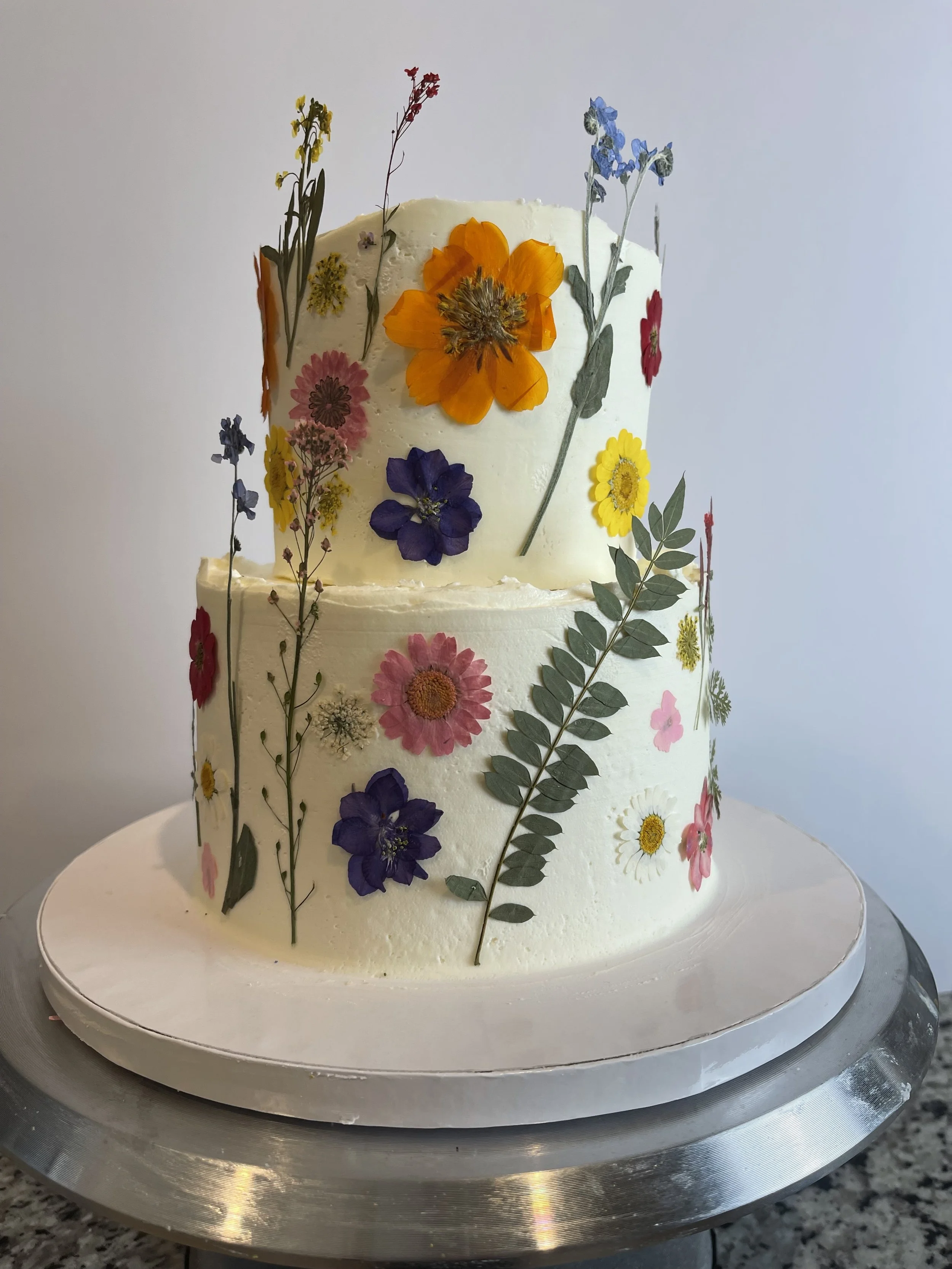 Two-tiered white cake decorated with edible pressed flowers and leaves, placed on a silver cake stand.