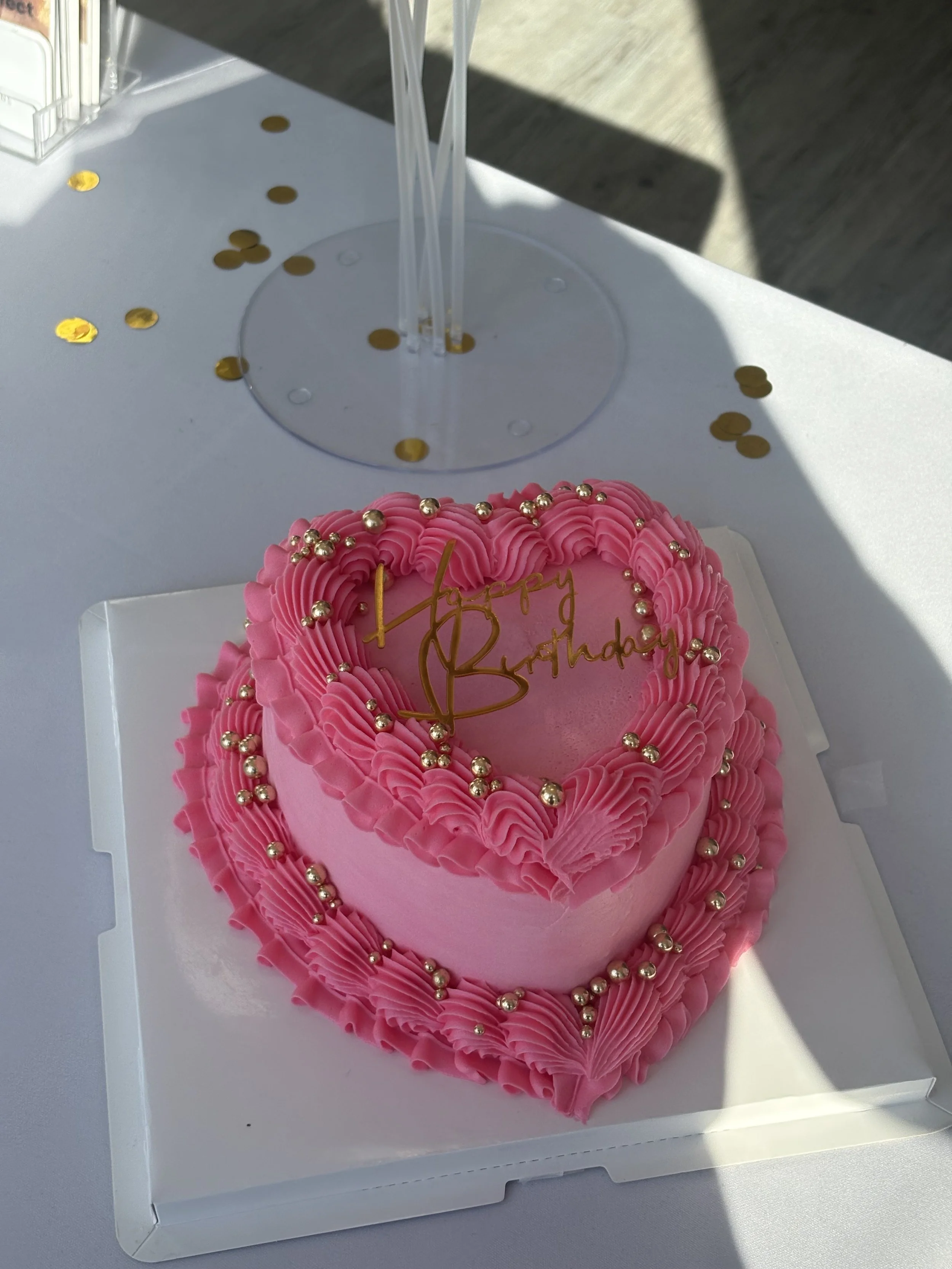 Pink heart-shaped birthday cake decorated with pink piped frosting and gold beads, with a gold "Happy Birthday" topper, placed on a white table with gold confetti and a clear plastic container in the background.