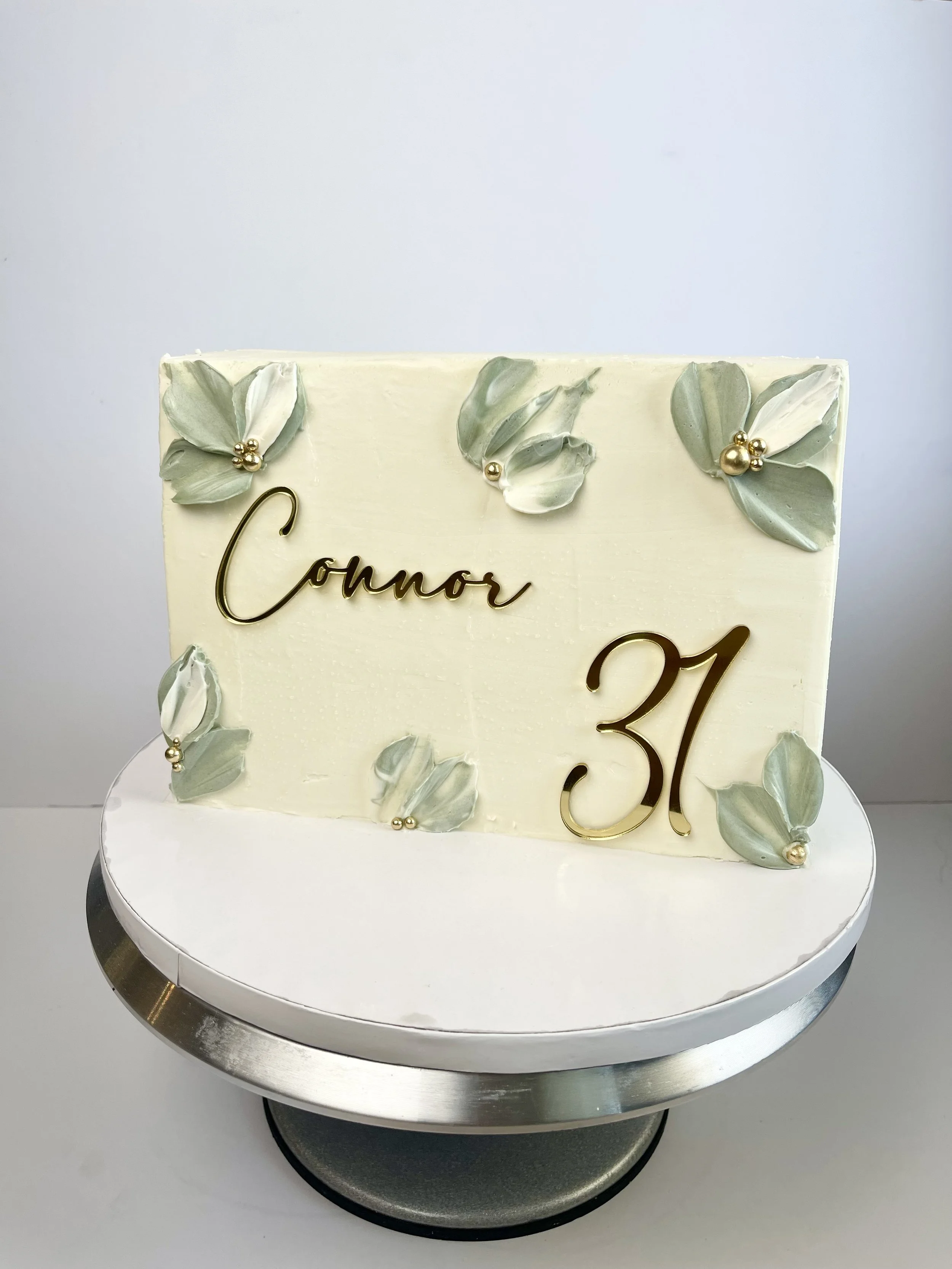 A white cake with green and white flower decorations, gold beads, the number 31 in gold, and the words 'Conor' on a silver cake stand.