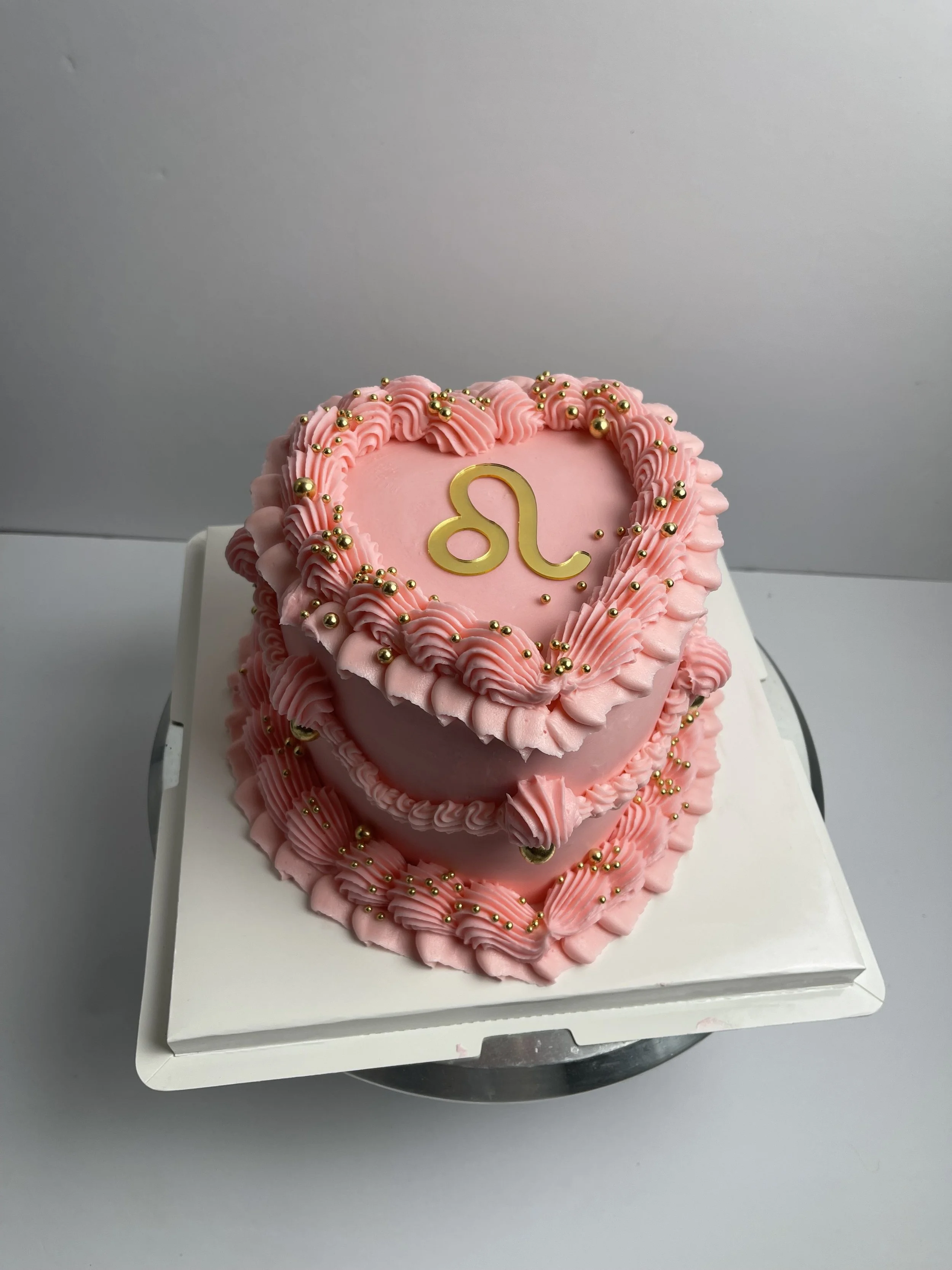 Pink heart-shaped cake with gold and pink decorative accents, featuring the Leo zodiac symbol on top.