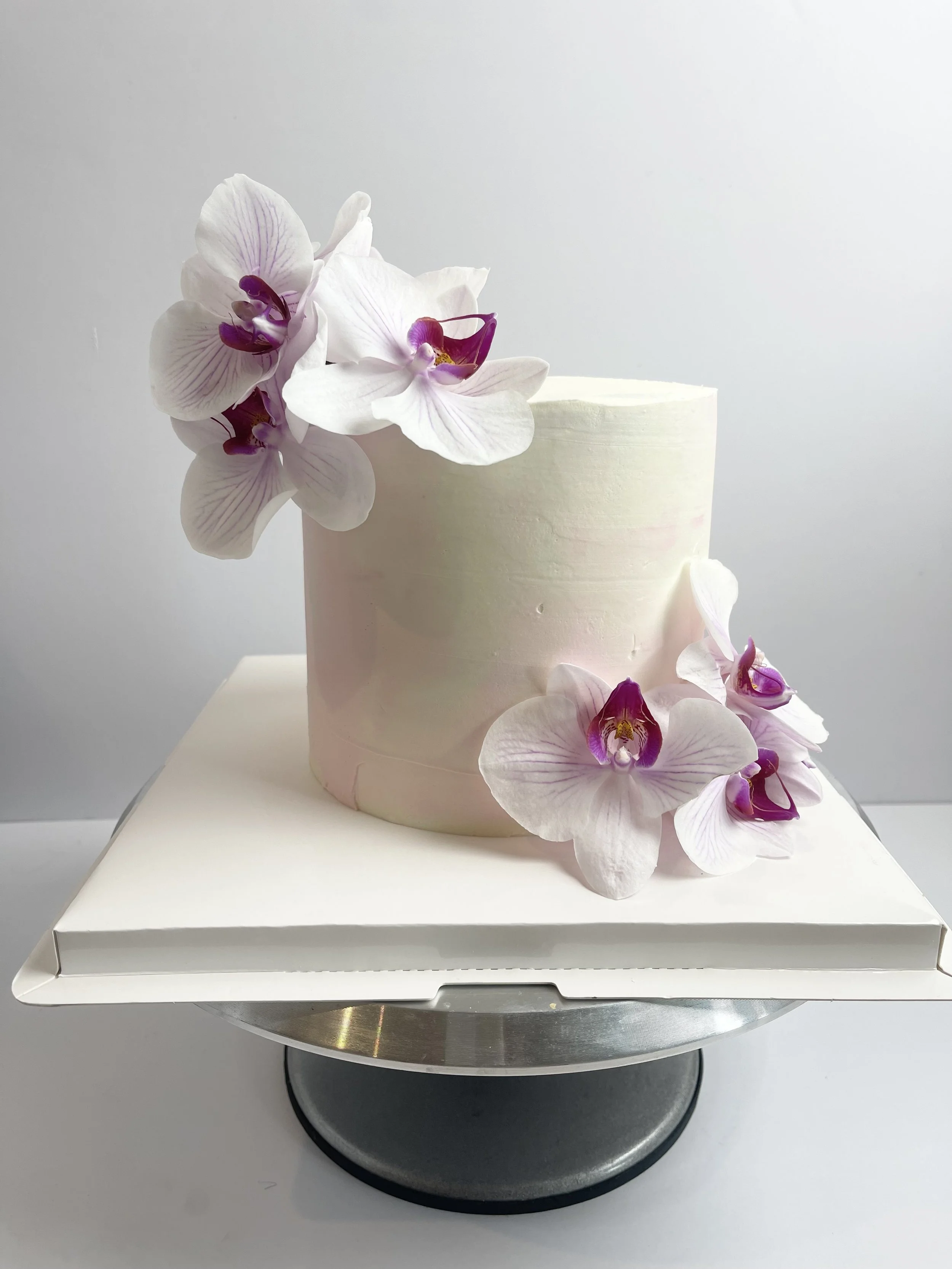 A white cylindrical cake decorated with purple and white orchids on top and on the side, placed on a white cake board and cake stand.