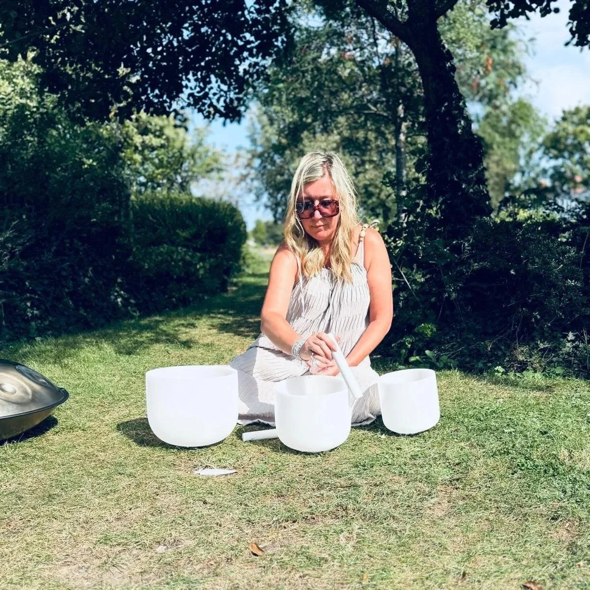 Woman sitting outdoors on grass, playing a crystal singing bowl set, surrounded by trees and bushes on a sunny day.