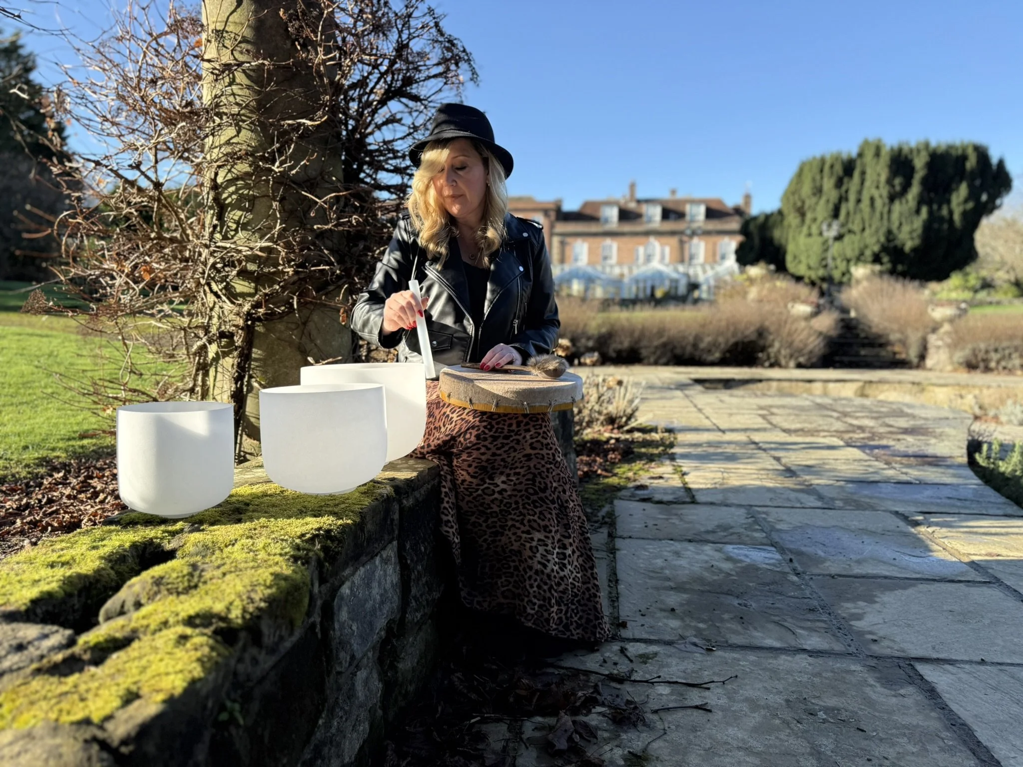 A woman with blonde hair, wearing a black hat and leather jacket, is sitting on a moss-covered stone wall outdoors on a sunny day, with three white singing bowls on the wall beside her, and a stone pathway extending into the distance with a large building and trees in the background.