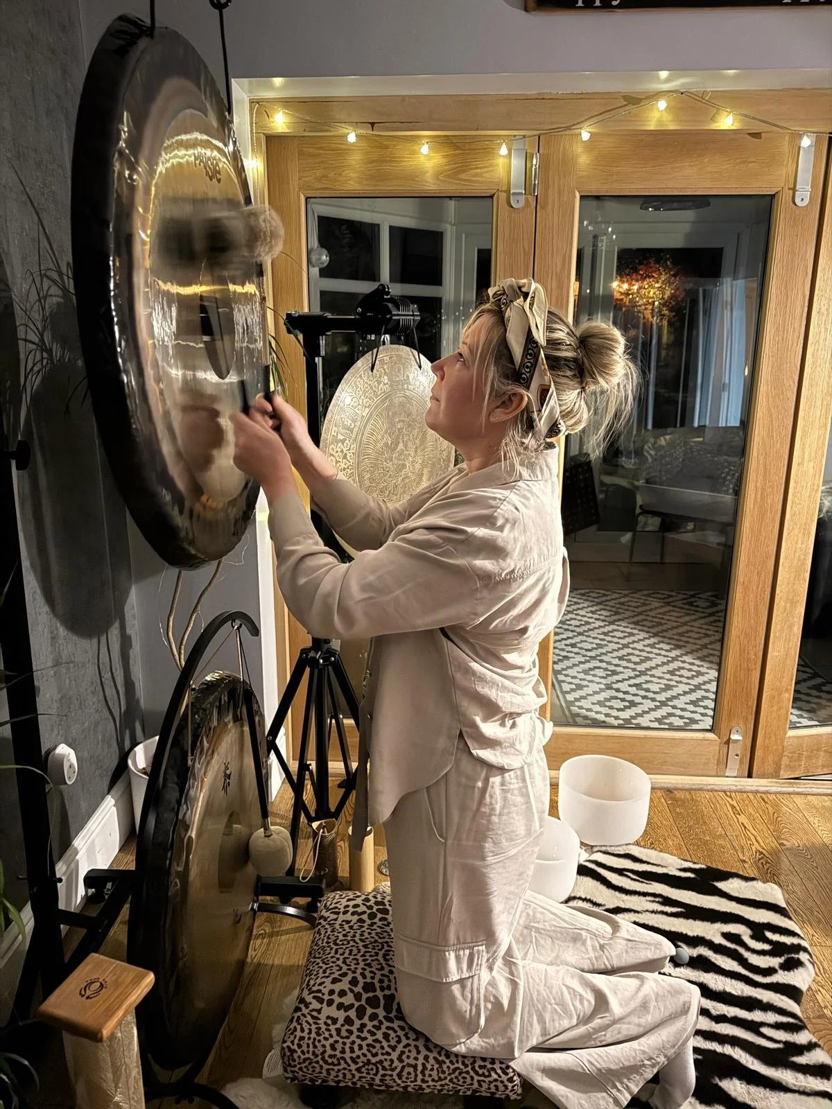 A woman kneeling on a leopard print cushion, playing a gong with a mallet in a cozy room decorated with string lights, a zebra print rug, and crystal singing bowls.