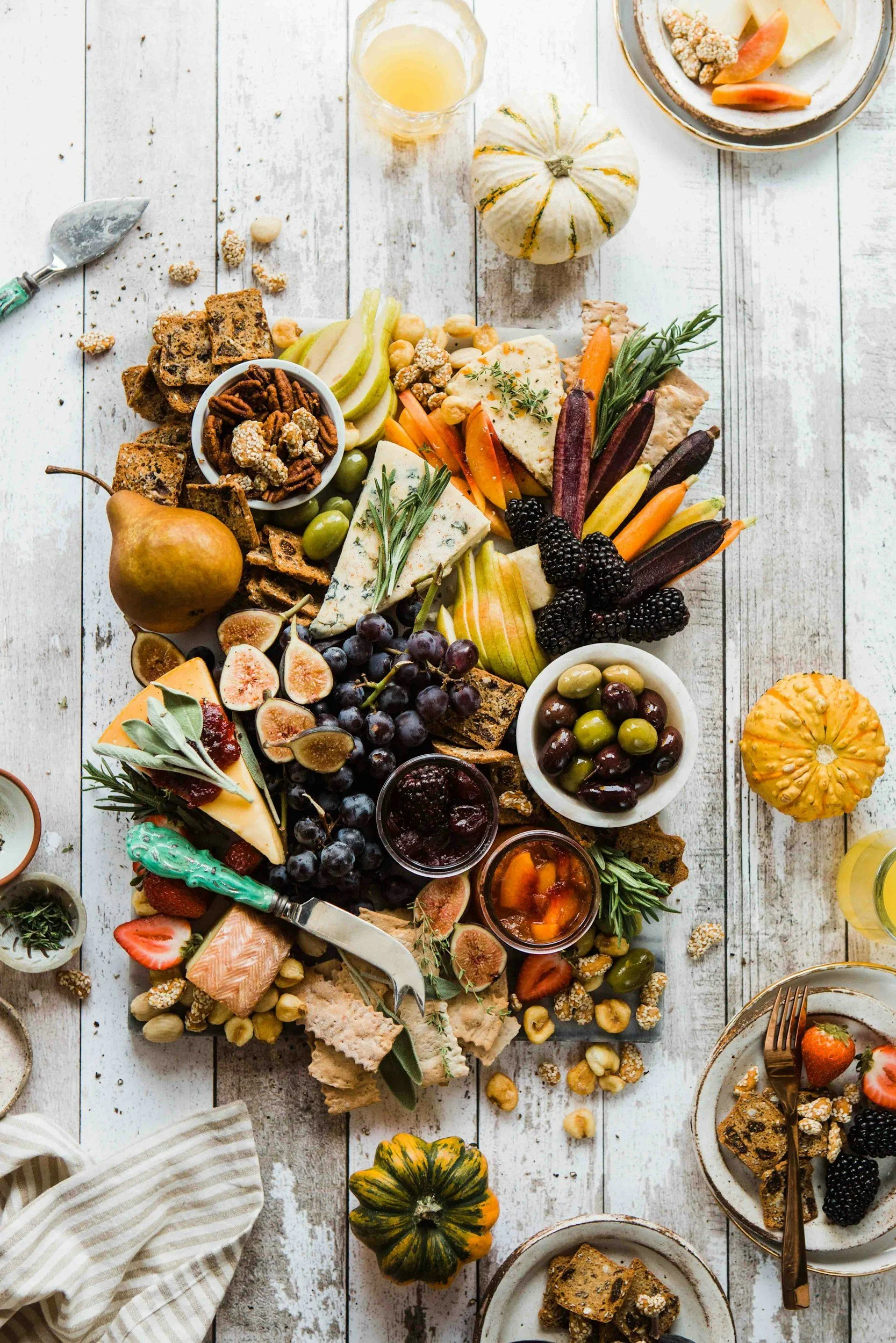 A festive charcuterie and cheese platter with fruits, nuts, and crackers on a rustic white wooden table, decorated with small pumpkins.