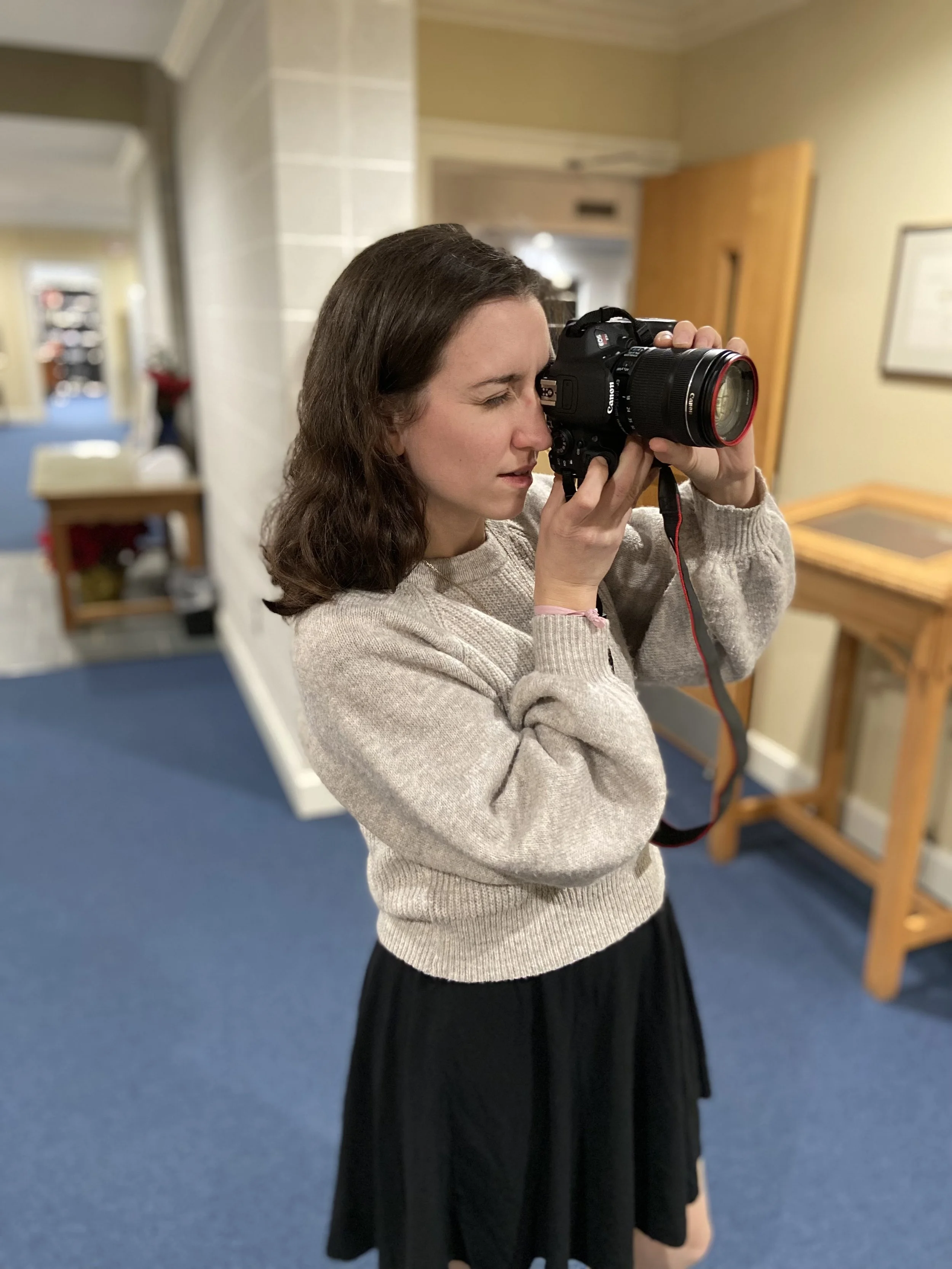 A woman taking a selfie in a mirror with a camera, with a beige sweater and a black skirt, in an indoor setting.