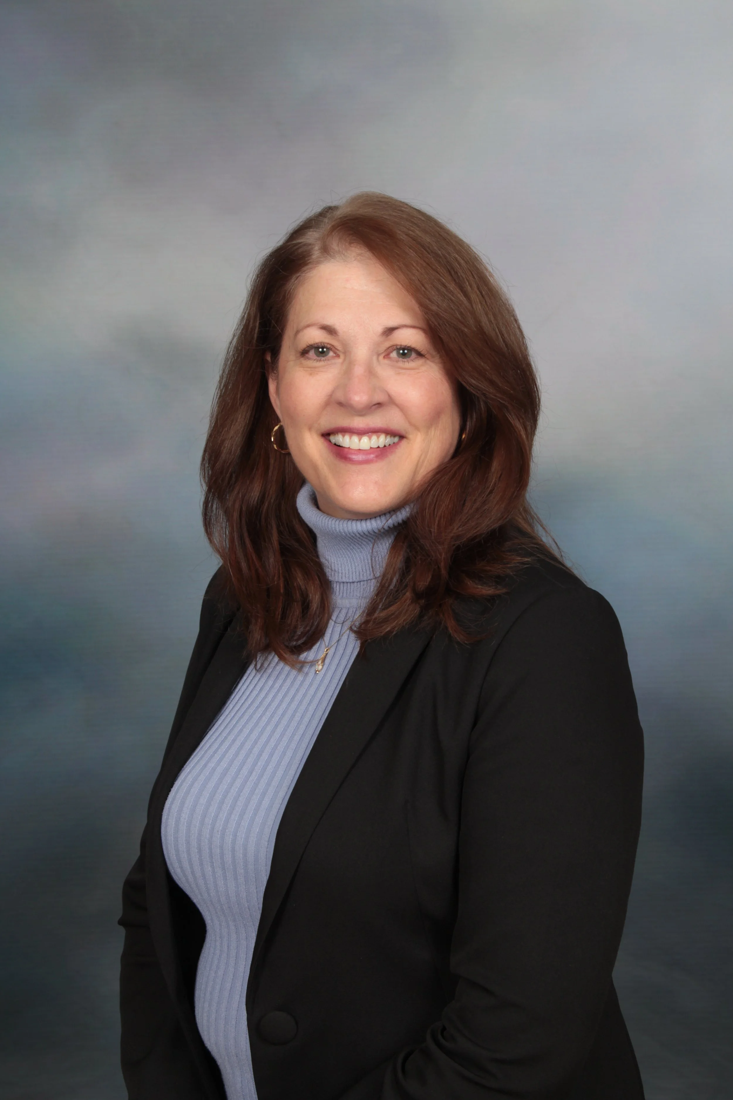 A professional woman with shoulder-length reddish-brown hair, wearing a black blazer over a light blue turtleneck, smiling against a cloudy sky background.