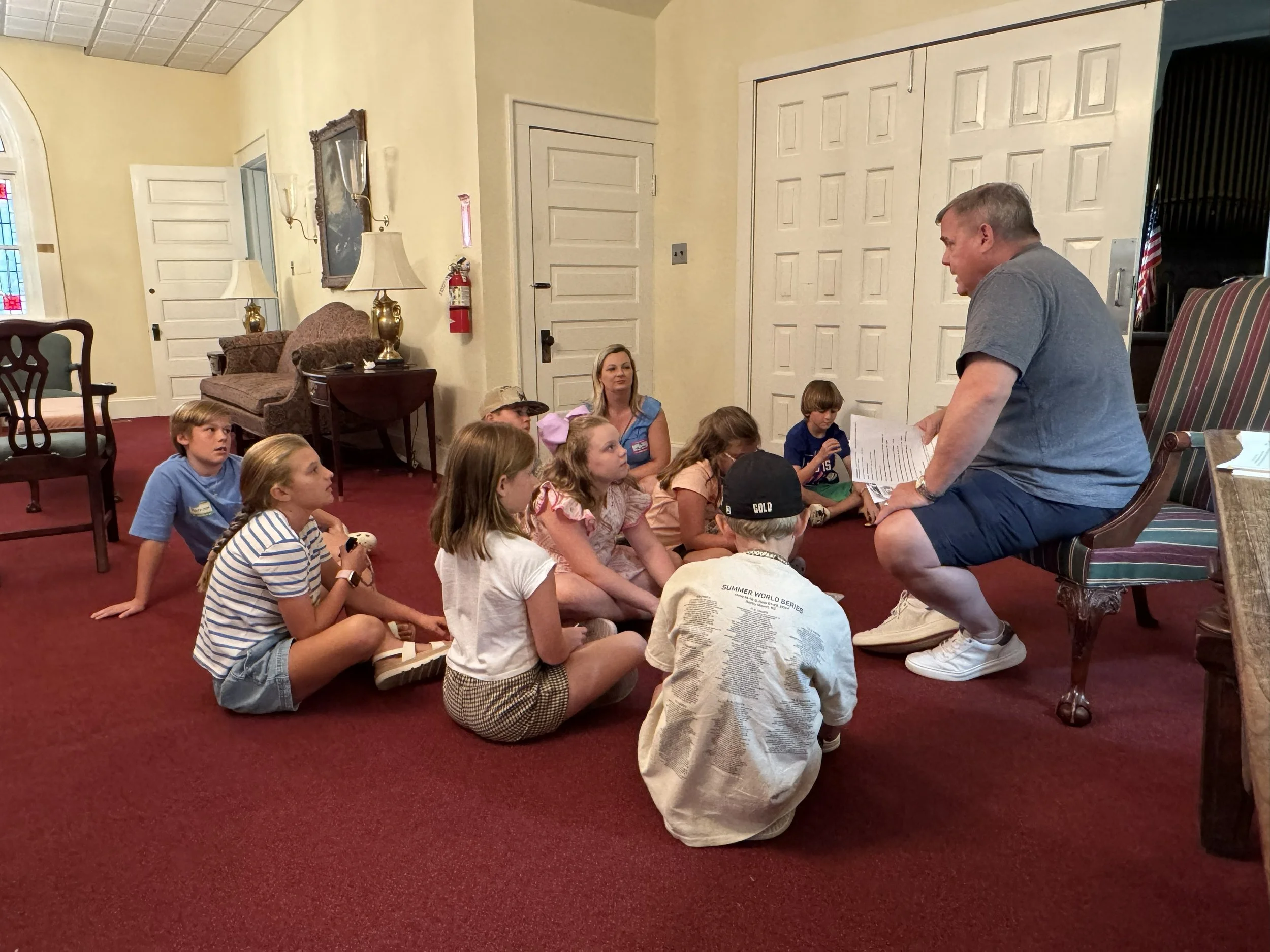 A man reading to a group of children sitting on the floor inside a room with red carpet and yellow walls.