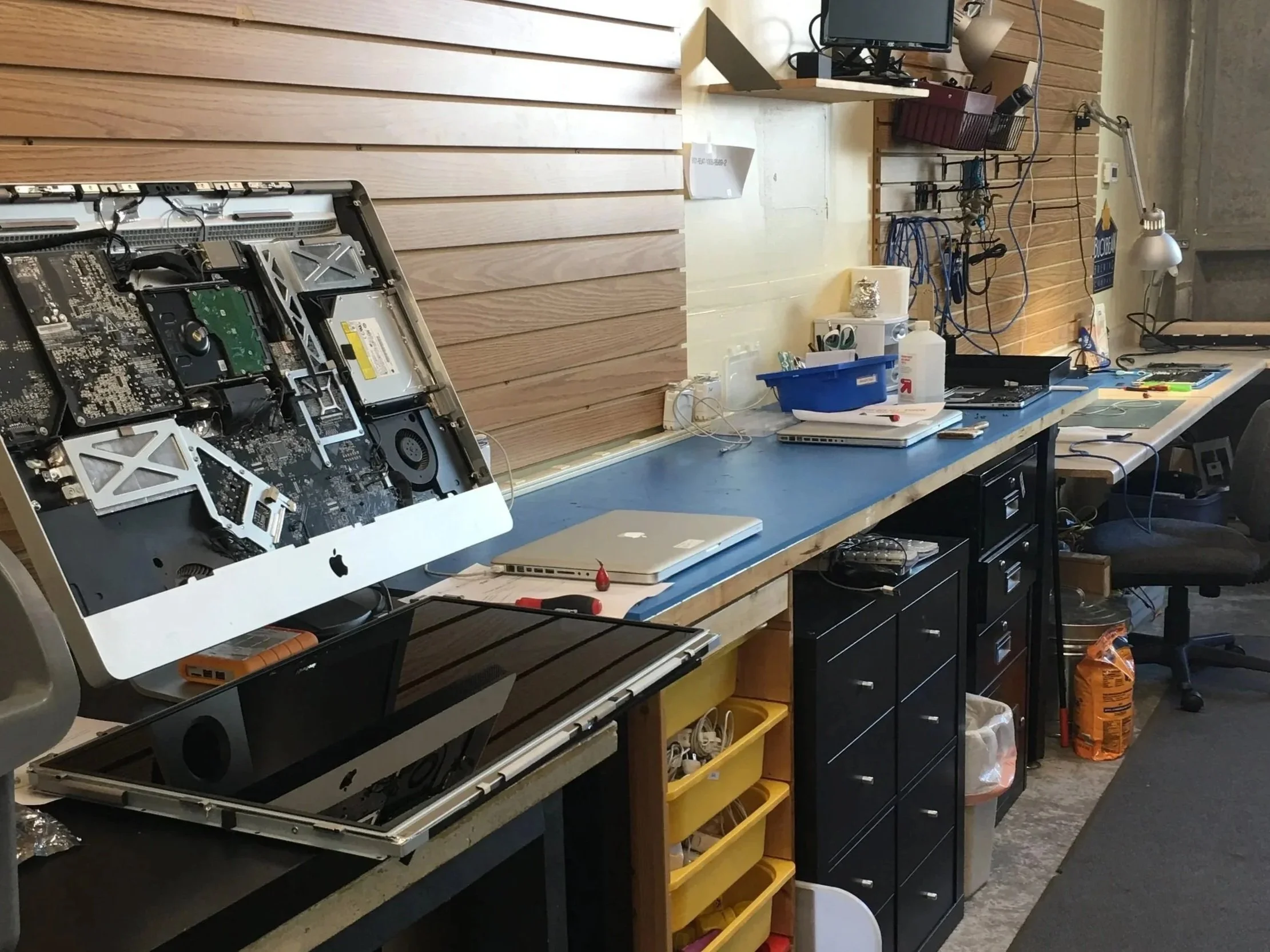 A workspace with an iMac computer disassembled, exposing internal components, on a workbench with various tools and electronic devices, against a wooden wall.