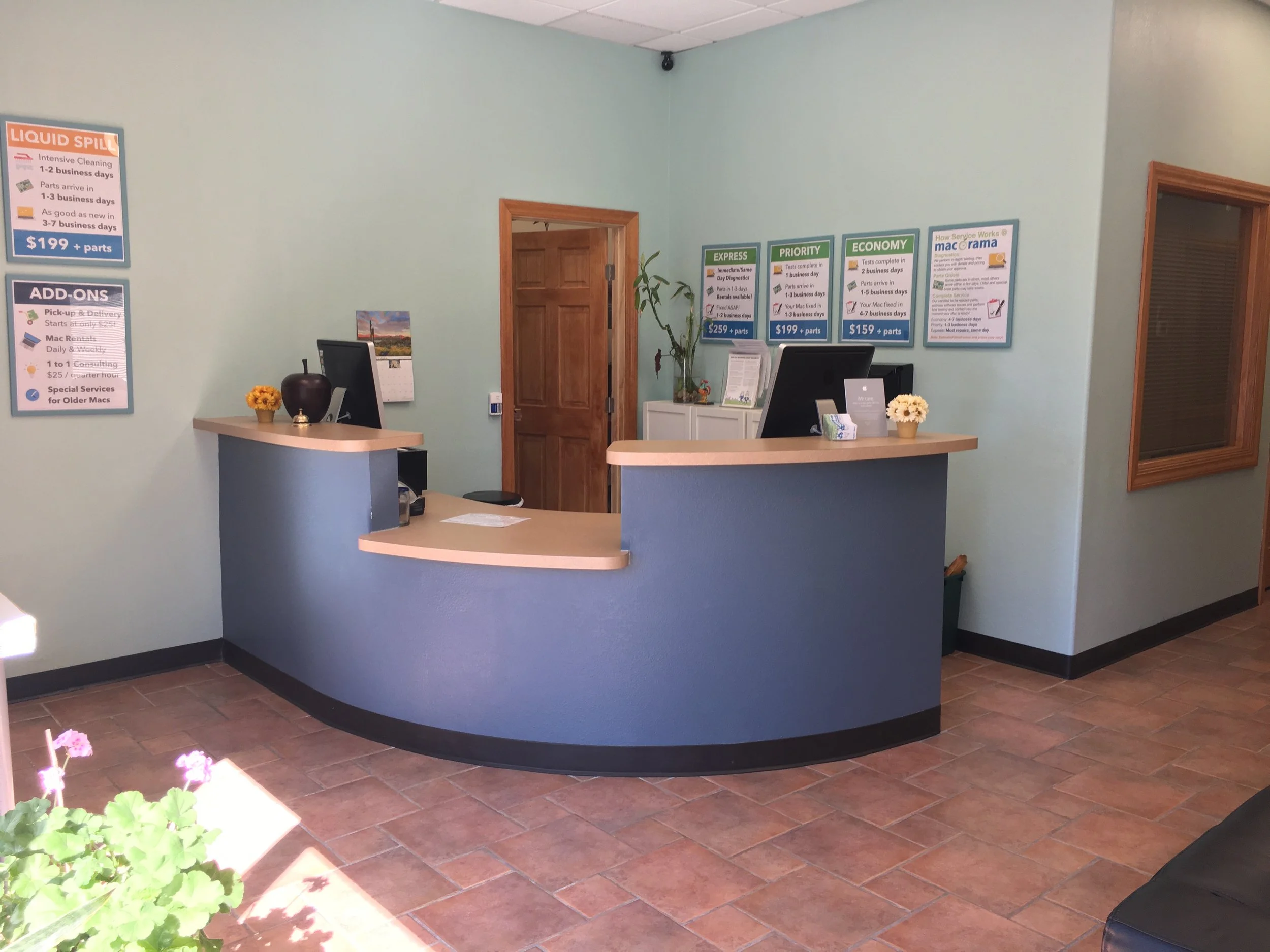 Reception desk with two computer monitors, a small vase with flowers, and promotional posters on the wall behind the desk in a car service center.