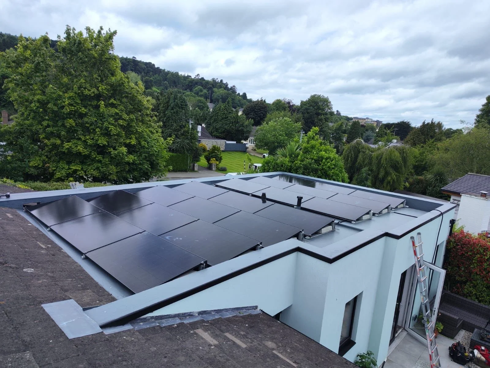 Solar panels installed on the flat roof of a modern white building, with green trees and cloudy sky in the background.