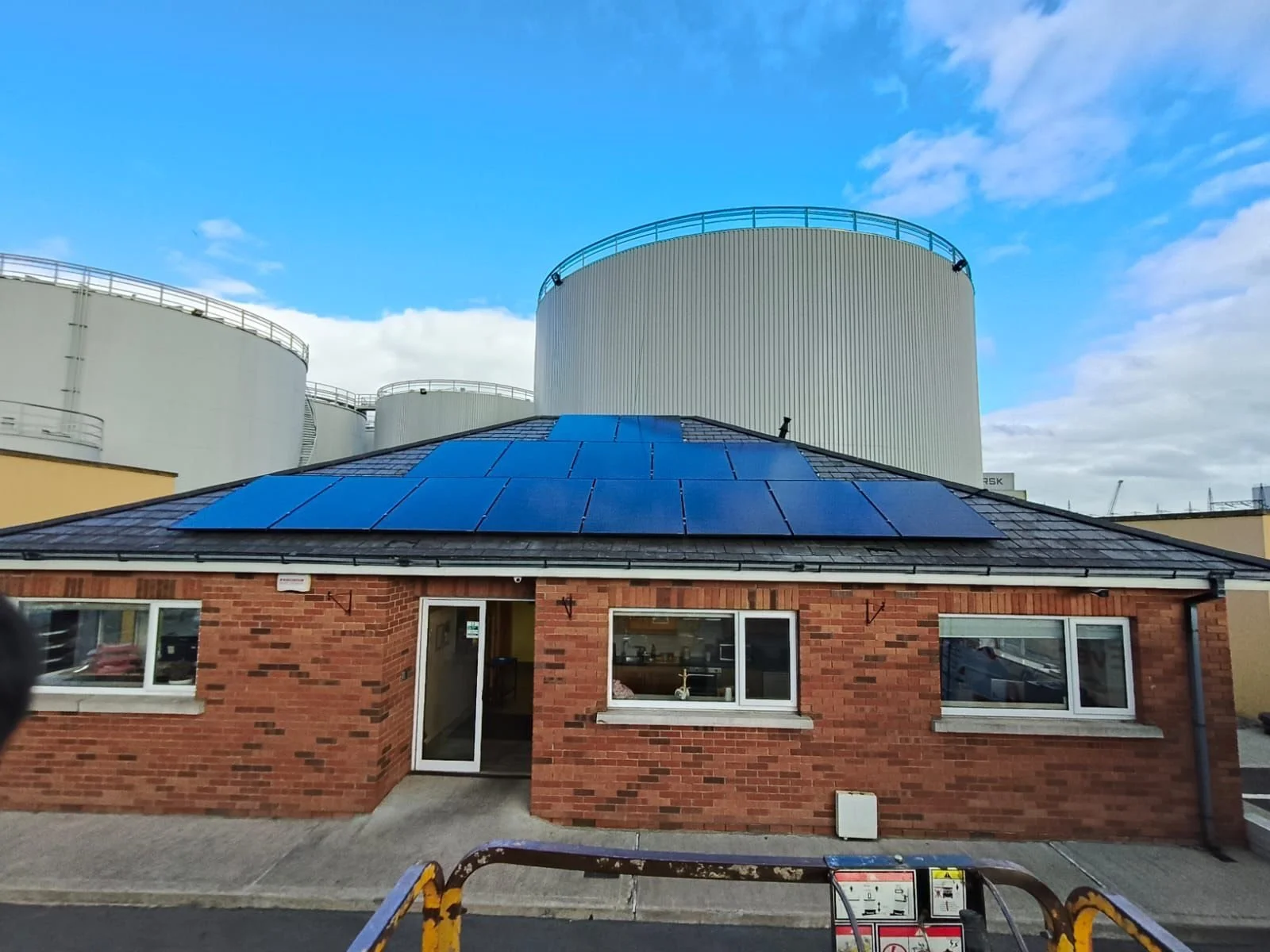 A small brick building with solar panels on the roof, with large industrial tanks behind it and a partly cloudy sky overhead.