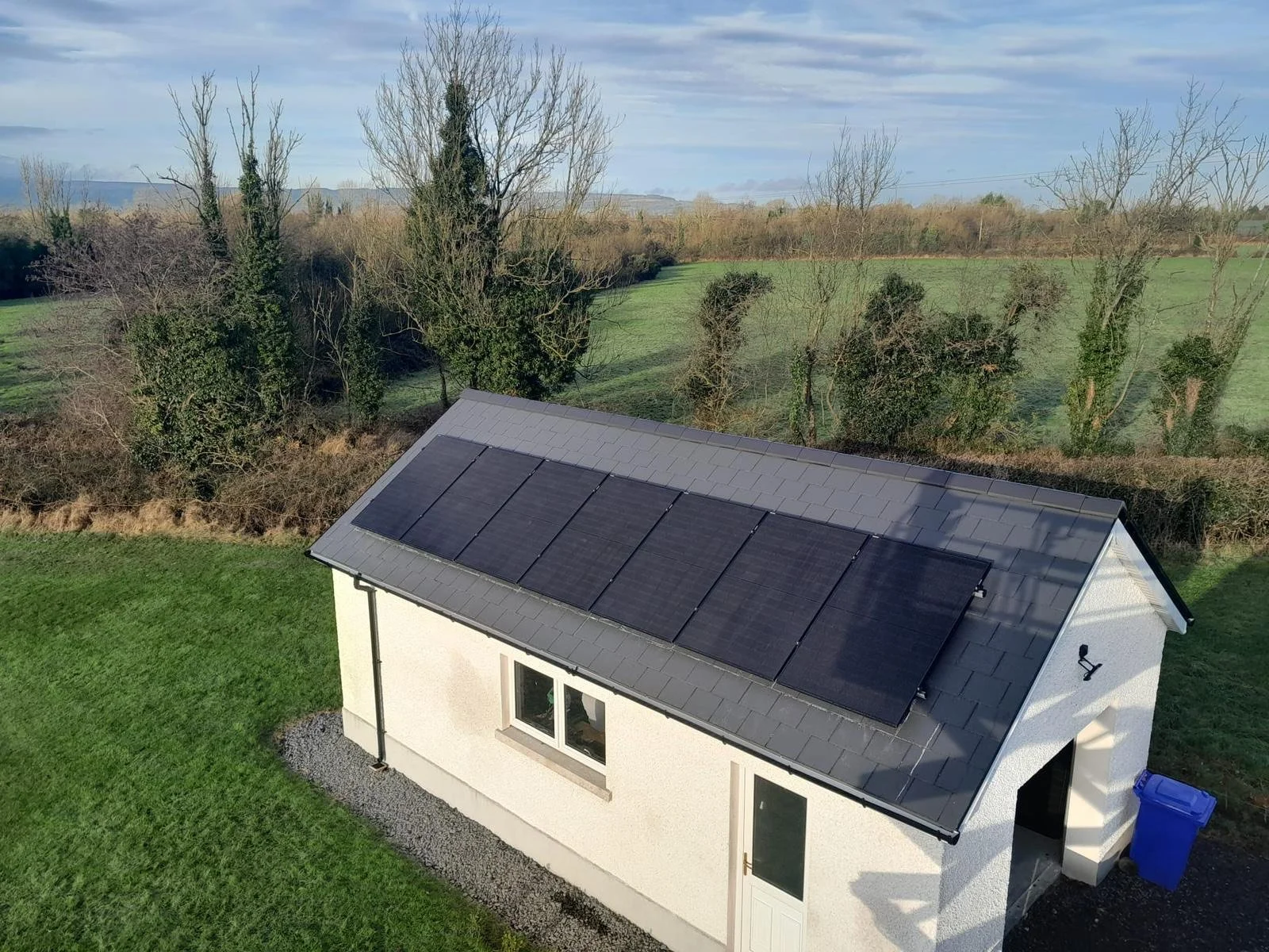 Small white house with solar panels on the roof, a window, a door, and a blue trash bin outside, set in a grassy area with trees and fields in the background.