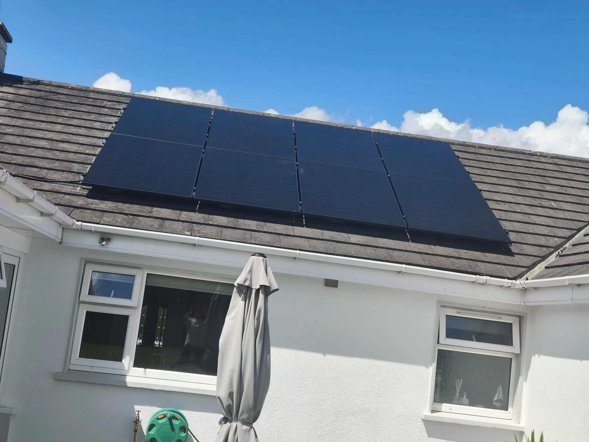 Solar panels installed on the roof of a white house with partial windows and garden furniture below on a sunny day with blue sky and clouds.