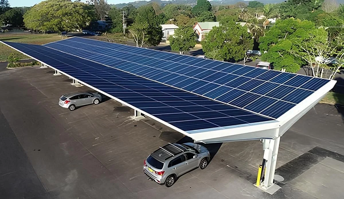 Solar panels installed on a parking lot cover a few parked cars with trees and buildings in the background.