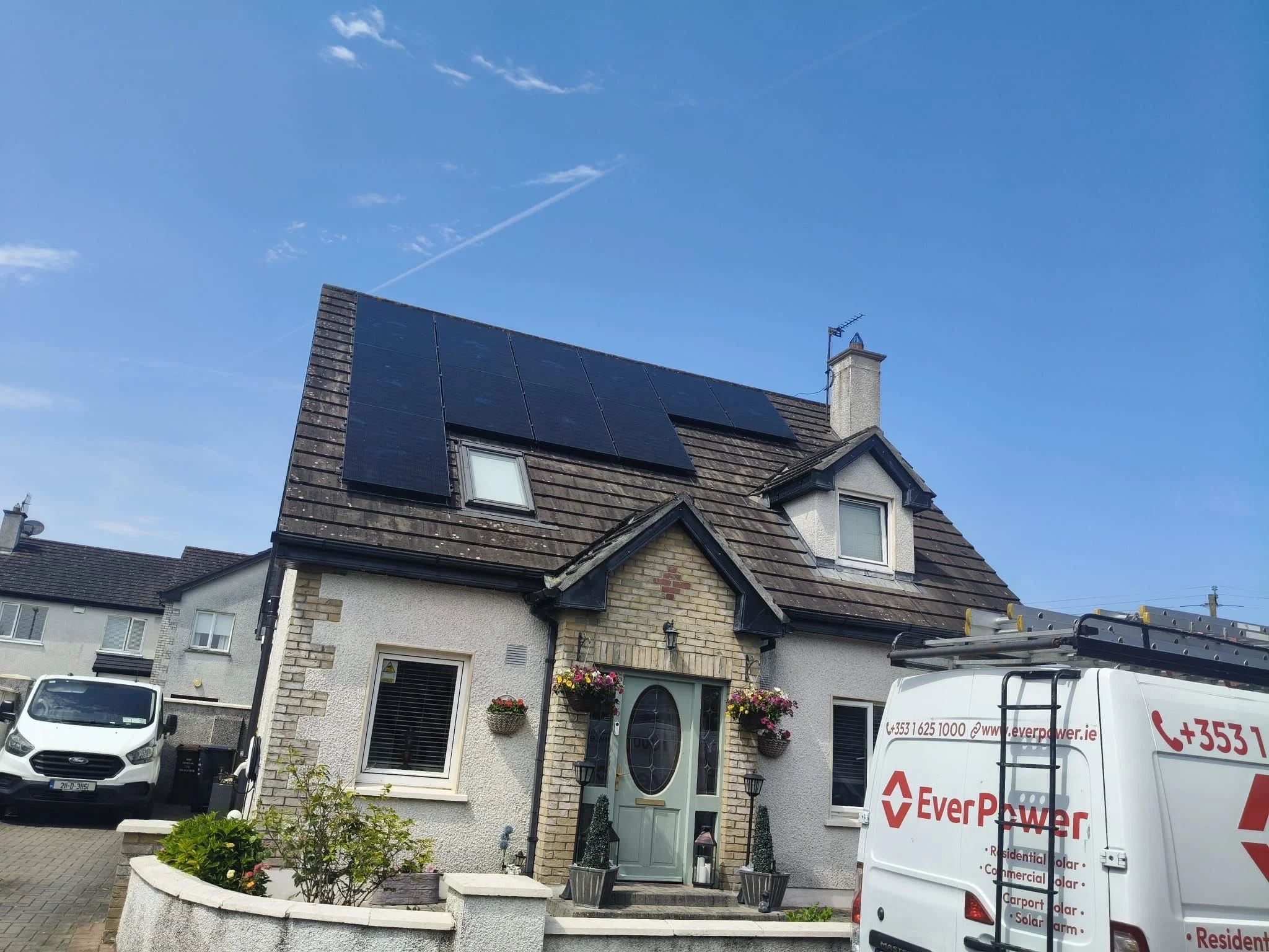A house with solar panels installed on the roof, featuring a front door decorated with hanging flower baskets and a small garden. There is a white van with company branding and a ladder parked nearby.