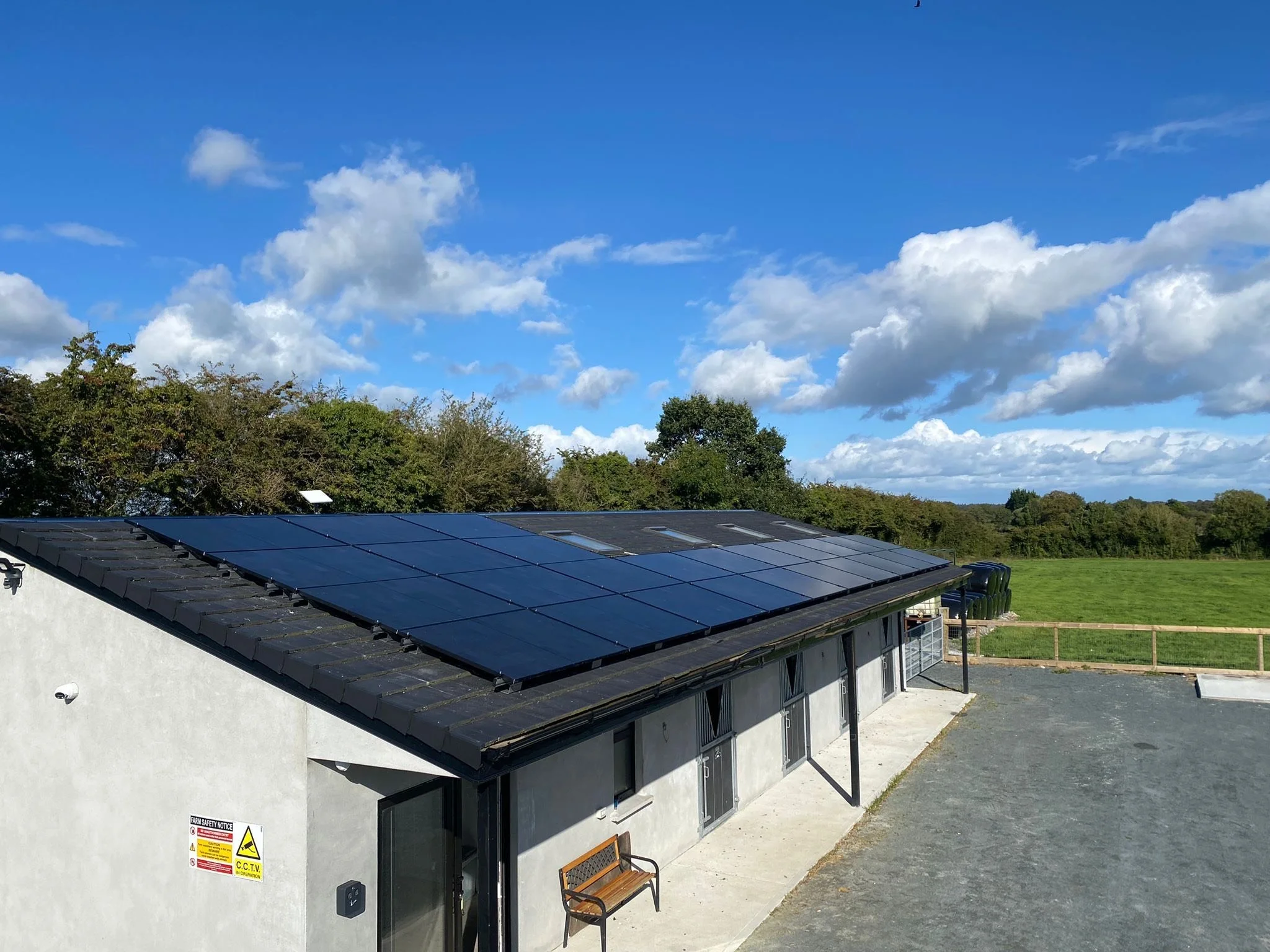 A building with solar panels on the roof, situated in a rural area with a grassy field and trees in the background, under a partly cloudy blue sky.